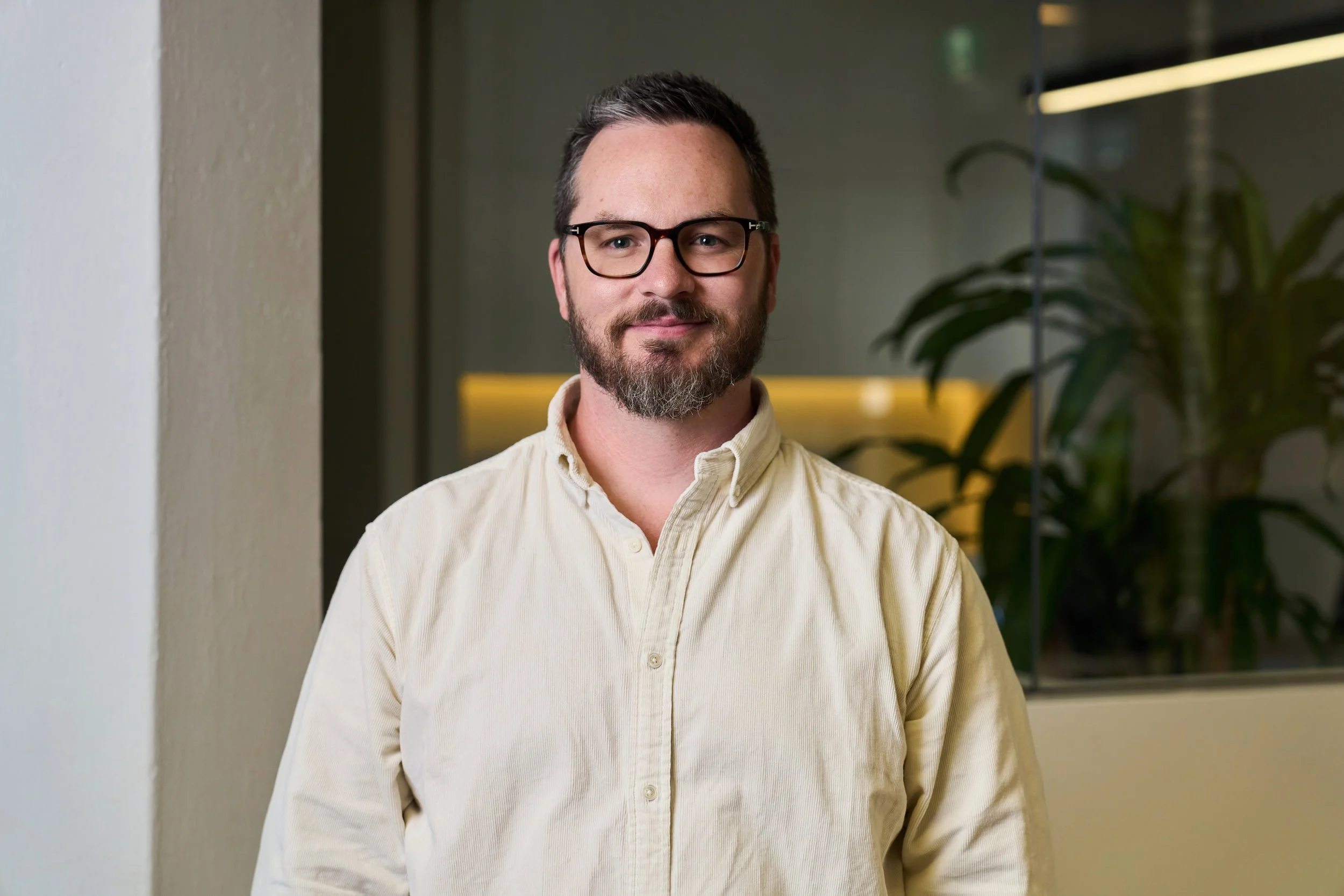 A man with glasses, short dark hair, and a beard, wearing a black t-shirt and a maroon hoodie, sitting outdoors against a cloudy sky.