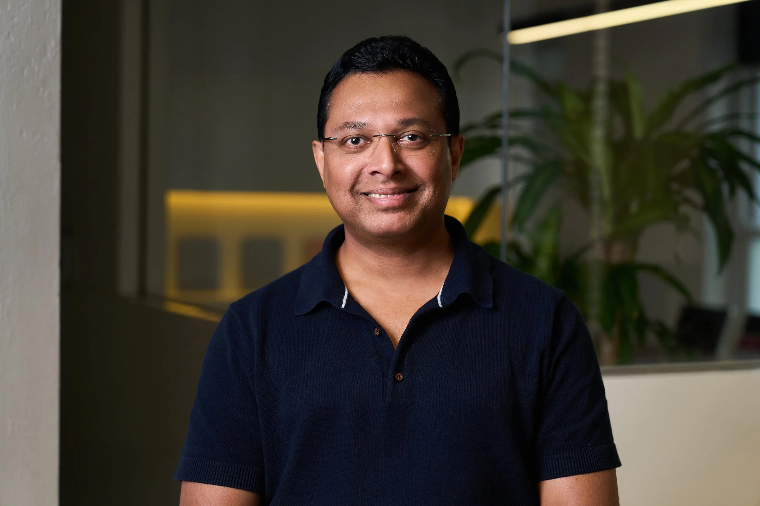 A professional man with glasses and dark hair, wearing a suit and tie, smiling against a grey background.