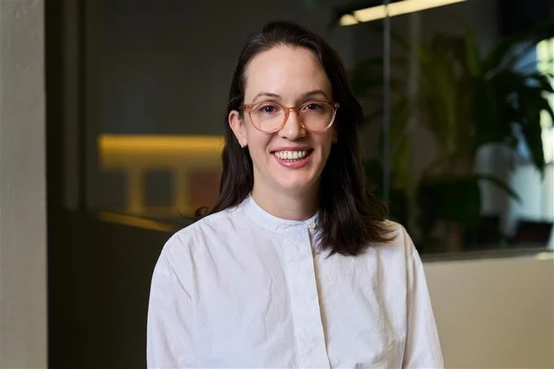 Person smiling, wearing glasses and a floral embroidered sweater, with a blurred whiteboard in the background.