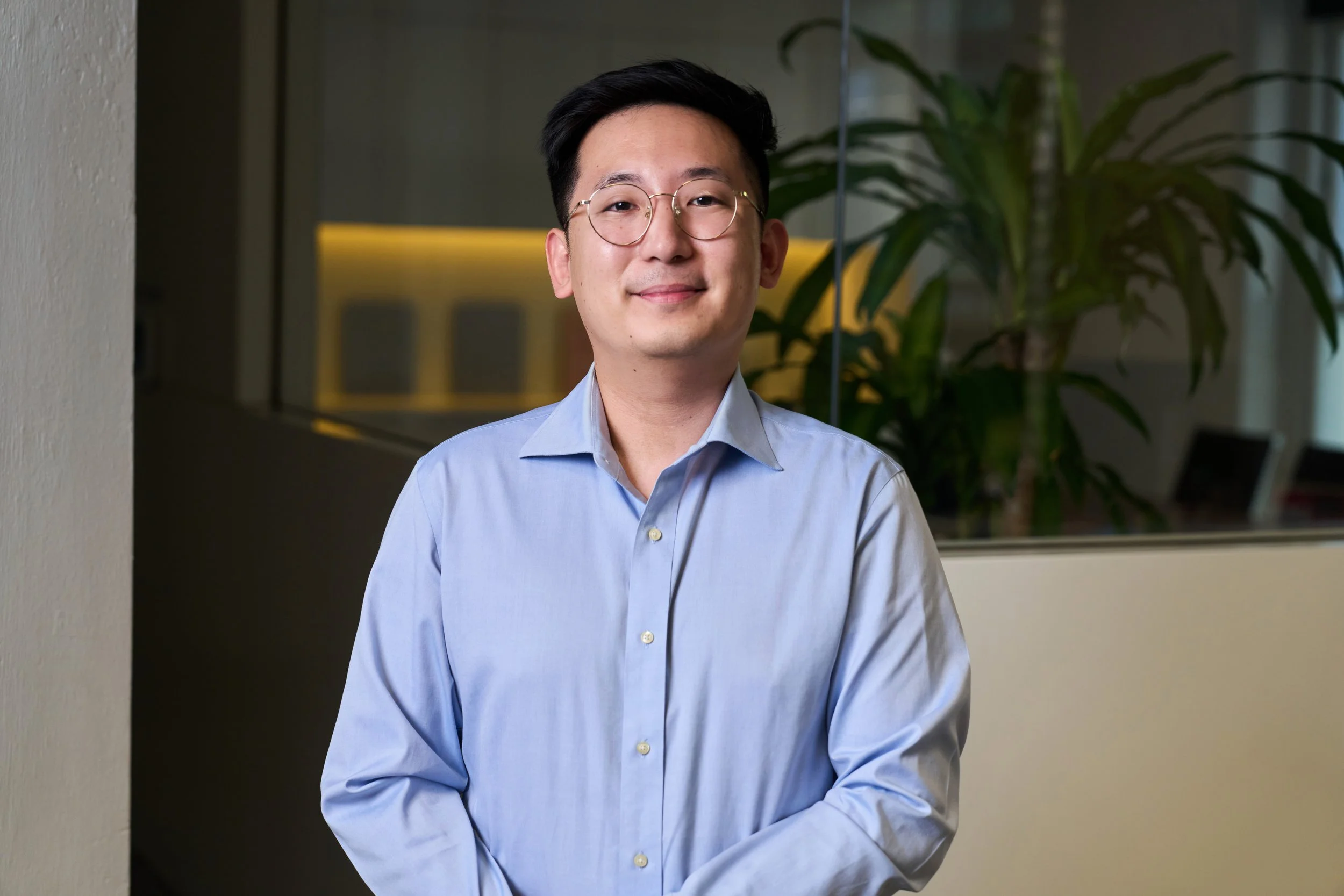 A person smiling and leaning against a tree outside with greenery in the background, wearing a dark green t-shirt.
