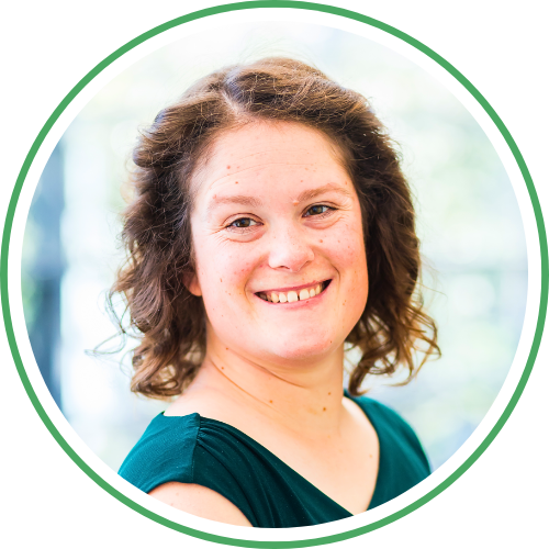 Ein Lächelndes Mädchen mit braunen, lockigen Haaren und einem dunkelgrünen Oberteil, vor einem unscharfen grauen und grünen Hintergrund.