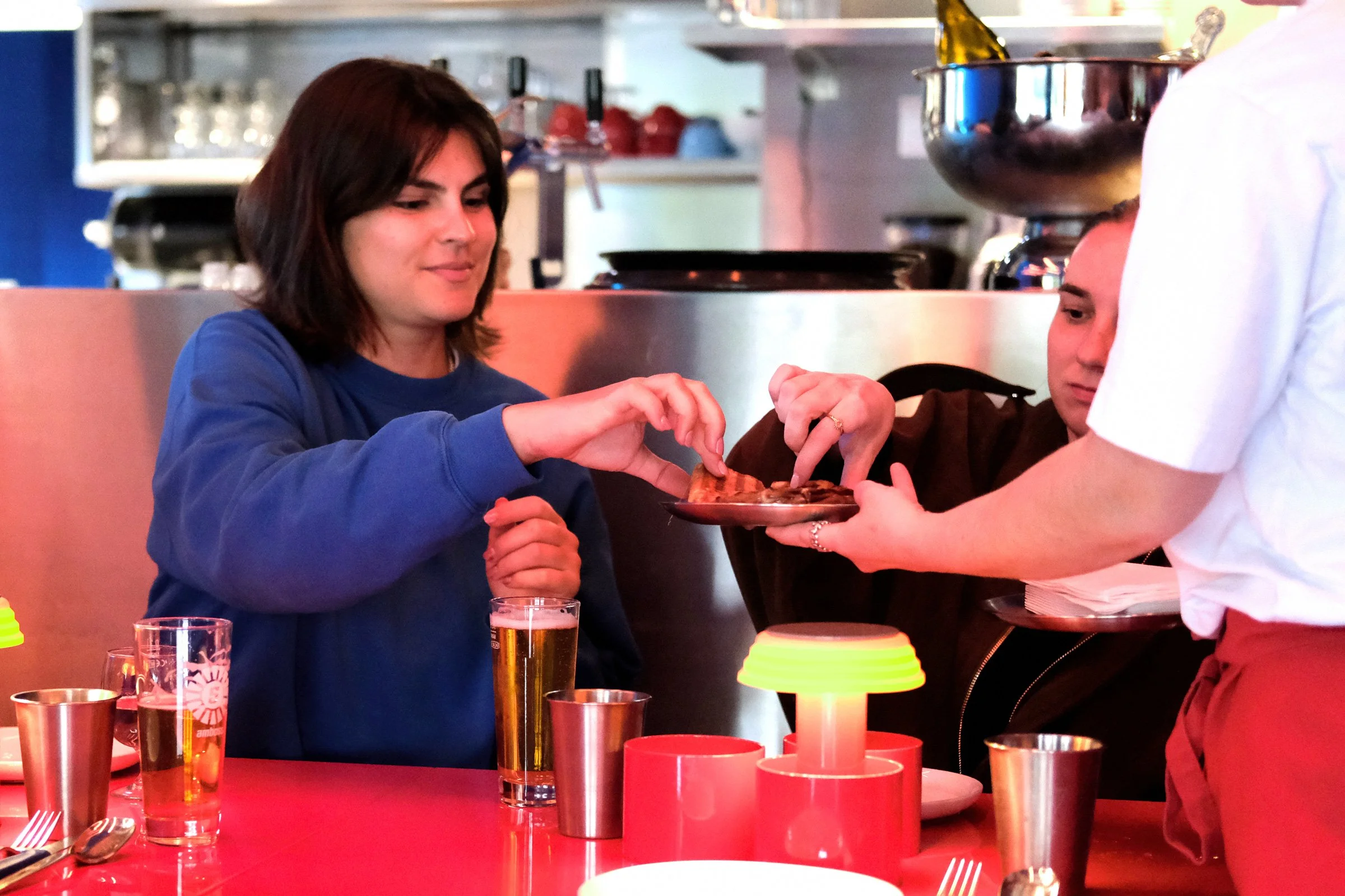 Eine Person in einem blauen Pullover erhält eine Pizza von einem Kellner in einem Restaurant. Es gibt Gläser mit Getränken und Kerzen auf dem roten Tisch.