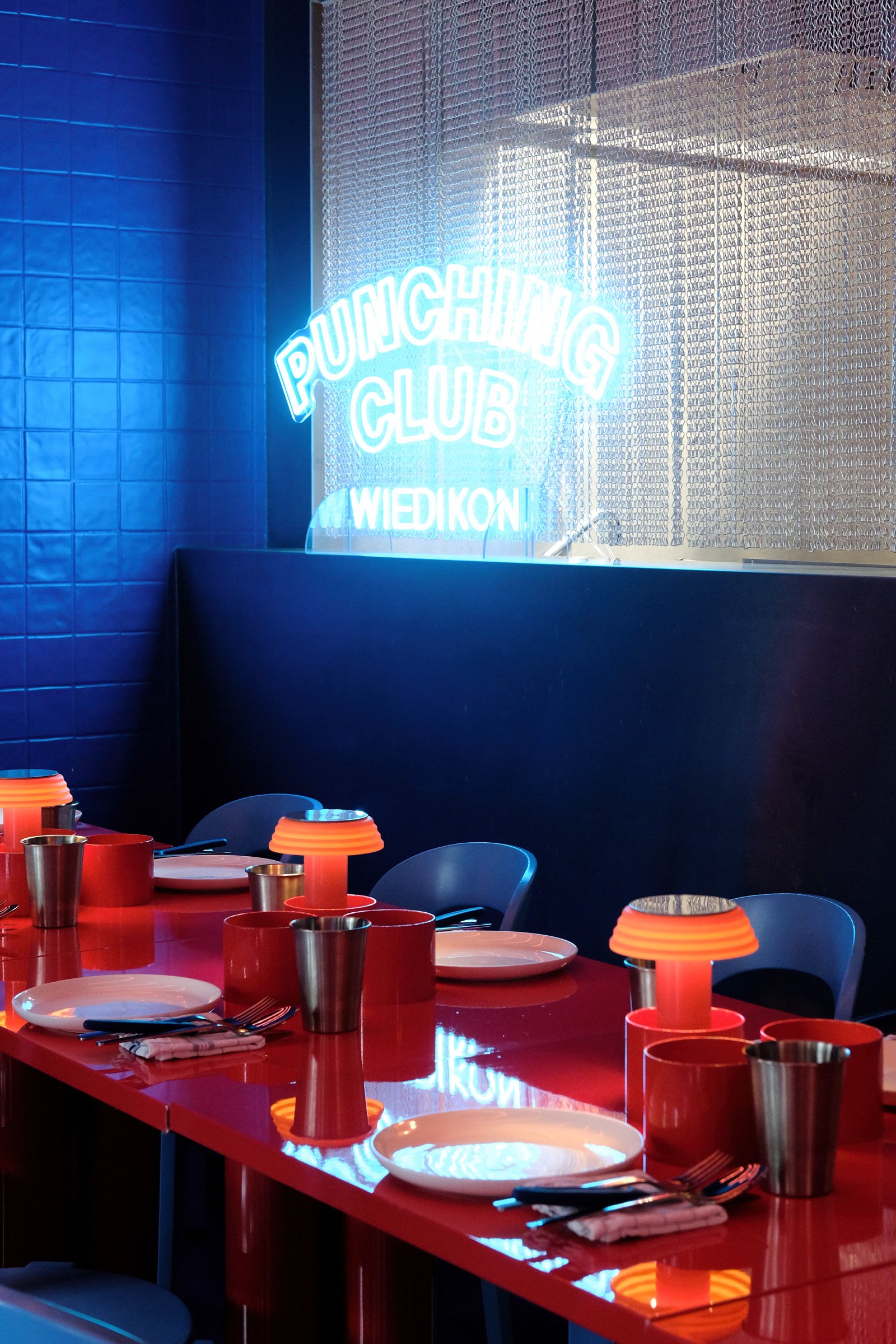 Ein modern dekorierter Raum mit einer roten Tafel, rot-weiße Tischtüchern, Kerzenlichtern und silbernen Bechern, im Hintergrund eine beleuchtete Neon-Schrift, die sagt 'PUNCHING CLUB WIEDIKON', alles in Blau- und Rottönen gestaltet.