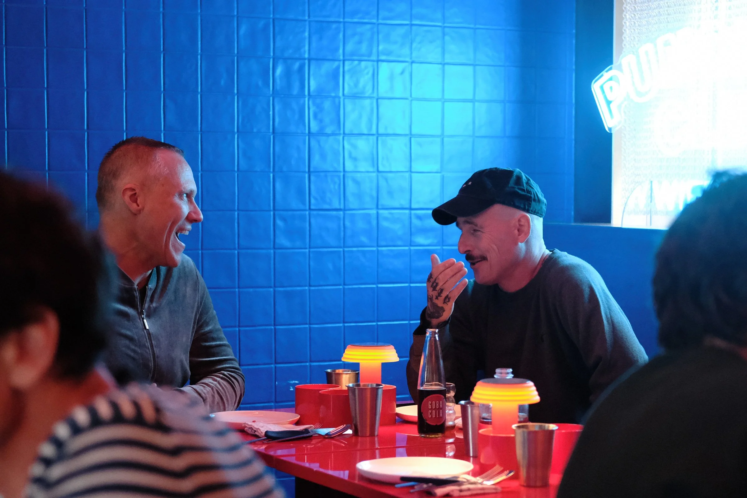 Zwei Männer sitzen an einem roten Tisch, lachen und unterhalten sich. Im Hintergrund blaue Fliesen und ein Neonlicht. Getränke und Teller auf dem Tisch.
