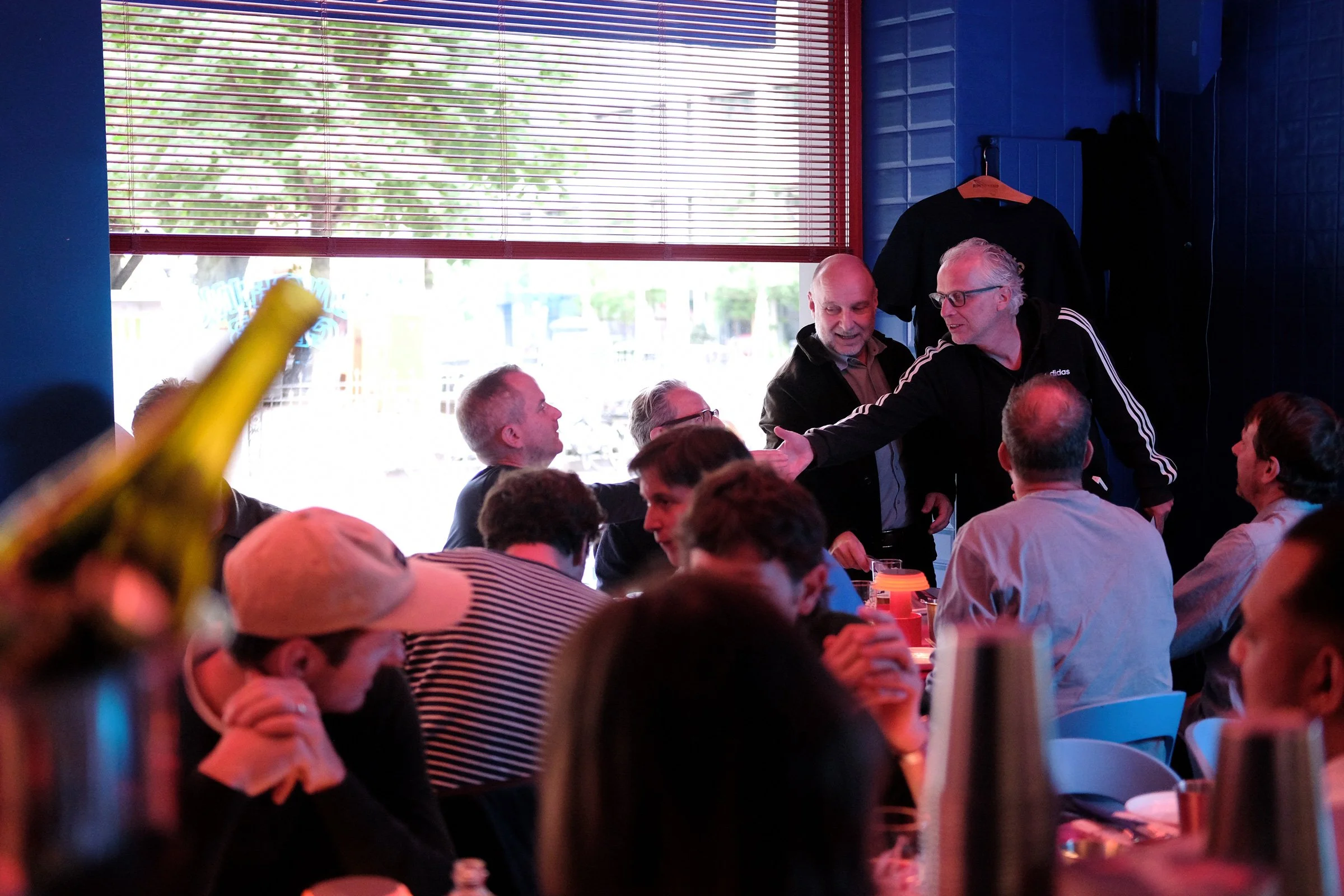 Menschen sitzen in einem hell erleuchteten Restaurant, einige unterhalten sich, während ein Mann mit grauen Haaren und Brille einem anderen die Hand schüttelt.