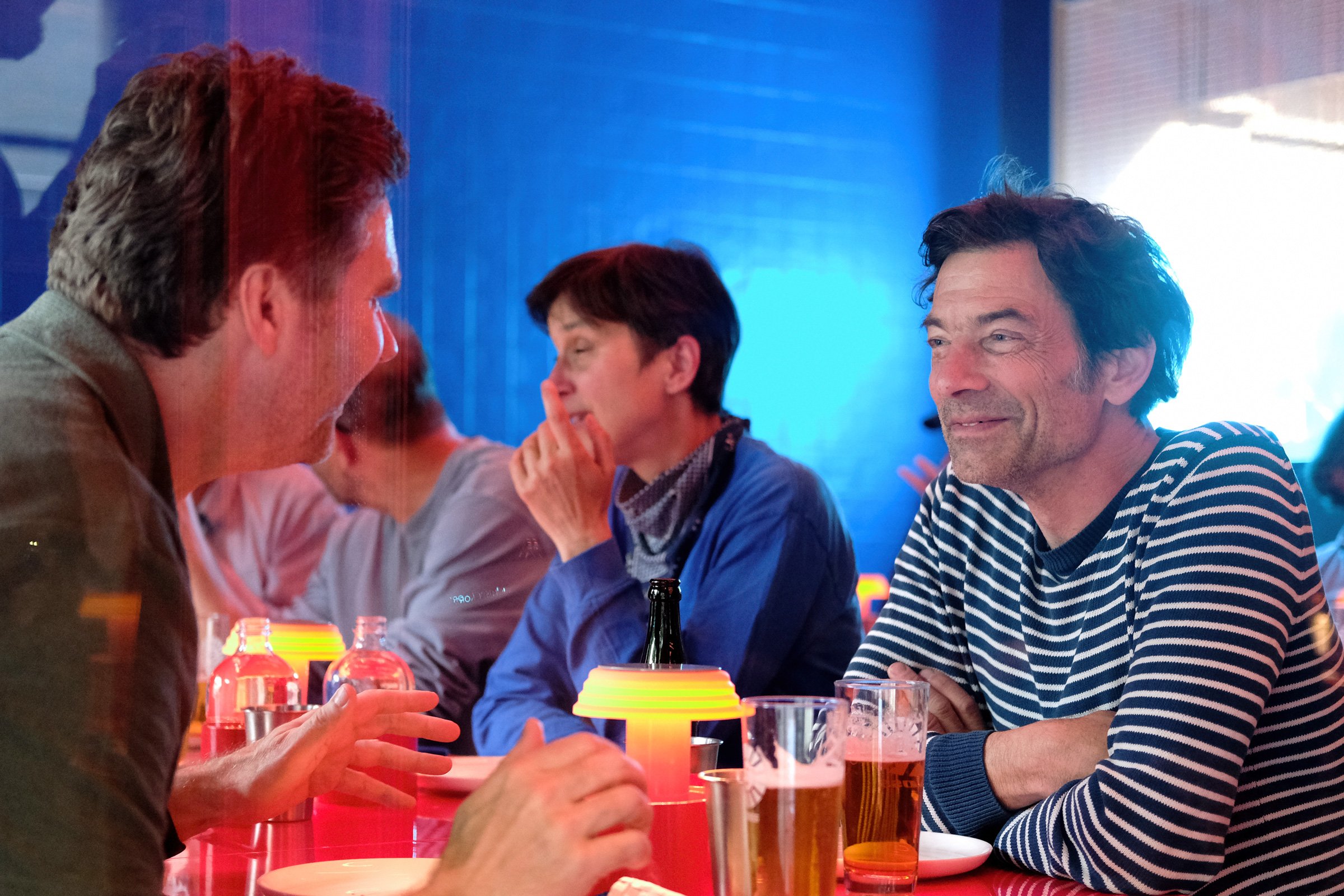 Menschen sitzen an einem Tisch in einer Bar oder einem Restaurant, trinken Bier und unterhalten sich. Im Hintergrund sind blaue Beleuchtungen und Gläser zu sehen.