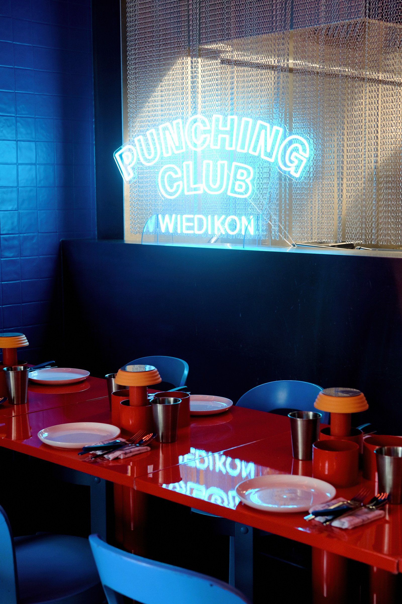 Blick auf einen roten Esstisch im Punching Club mit weißen Tellern, schwarzen Besteck, roten Trinkbechern, orangefarbenen Lampen und blauen Stühlen. Im Hintergrund ein beleuchtetes Neon-Schild mit der Aufschrift "Punching Club Wiedikon" und eine meta