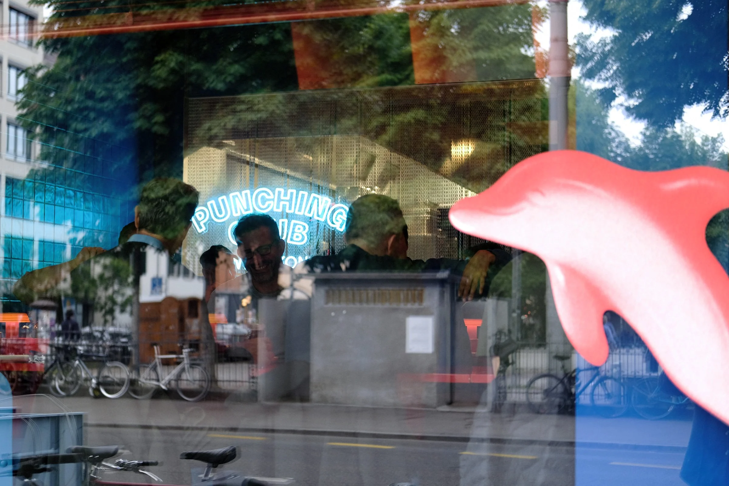 Reflexion eines Cafés mit einem Neonzeichen, Menschen und Freundschaften, draußen vor einem Geschäft in einer urbanen Umgebung, Fahrradständer und Fahrräder an der Straße.