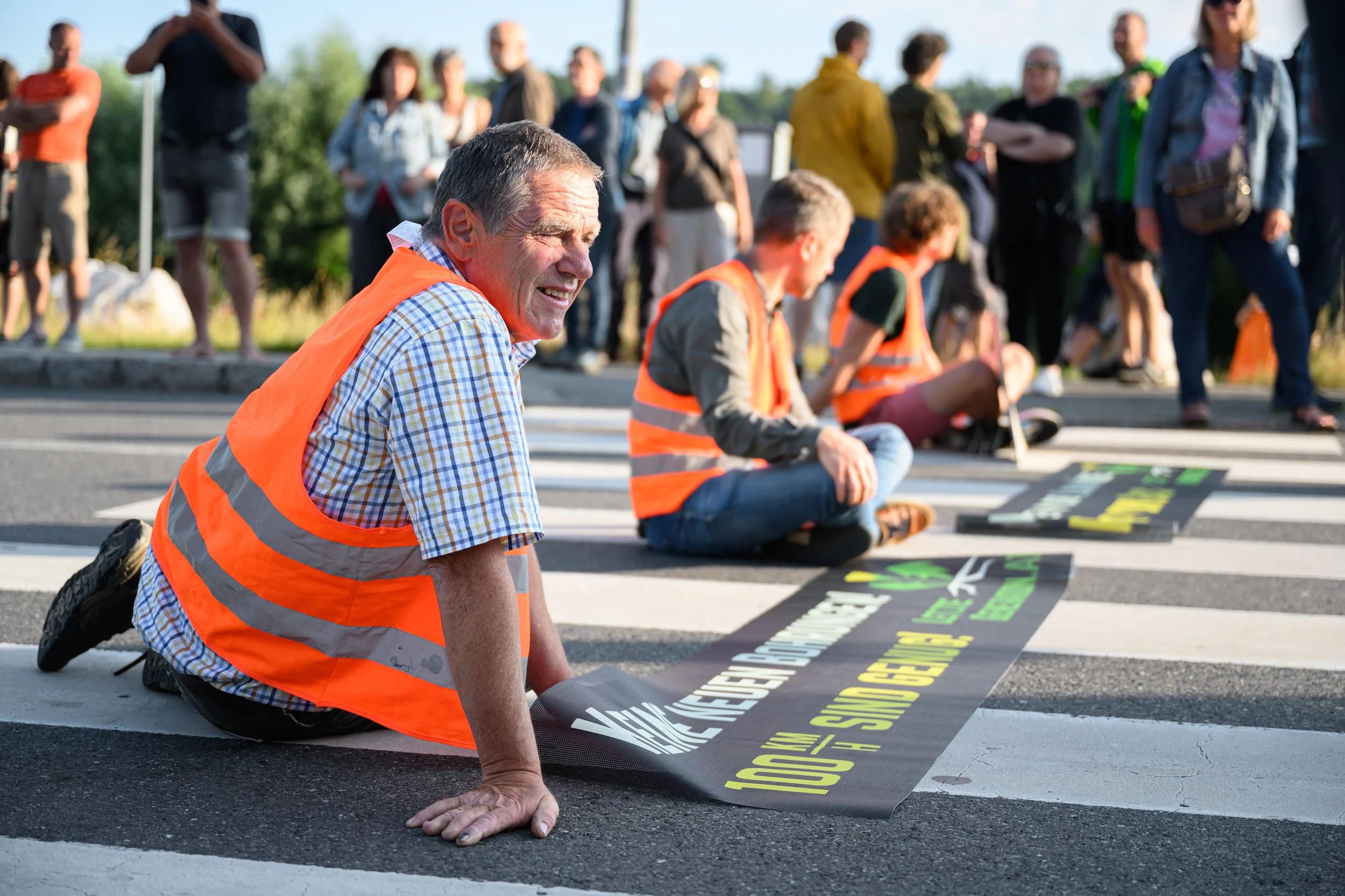 Ein älterer Herr hat sich mit zwei weiteren auf die Straße gesetzt. Im Hintergrund sieht man mehrere Menschen solidarisch dabei stehen.
