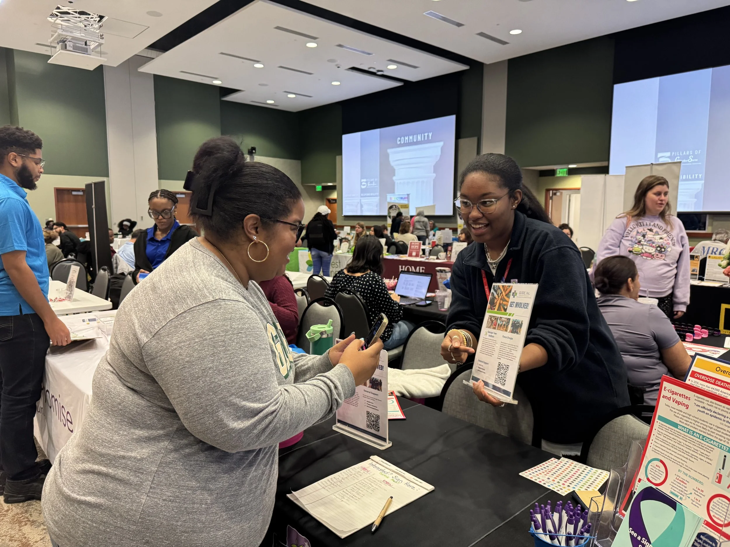 People interacting at health-related information booths in a conference room with large screens displaying community pillar graphics.