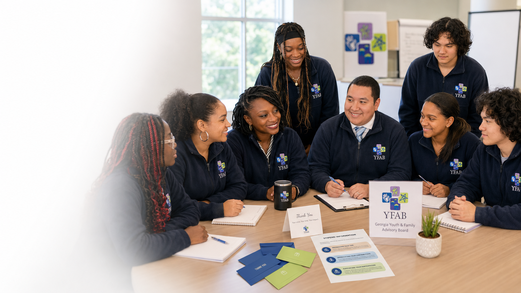 Group of young adults sitting around a table, engaging in a discussion, with a facilitator standing behind them. They are wearing matching navy blue jackets with the YFAB logo, in a bright room with large windows and informational materials on the table.