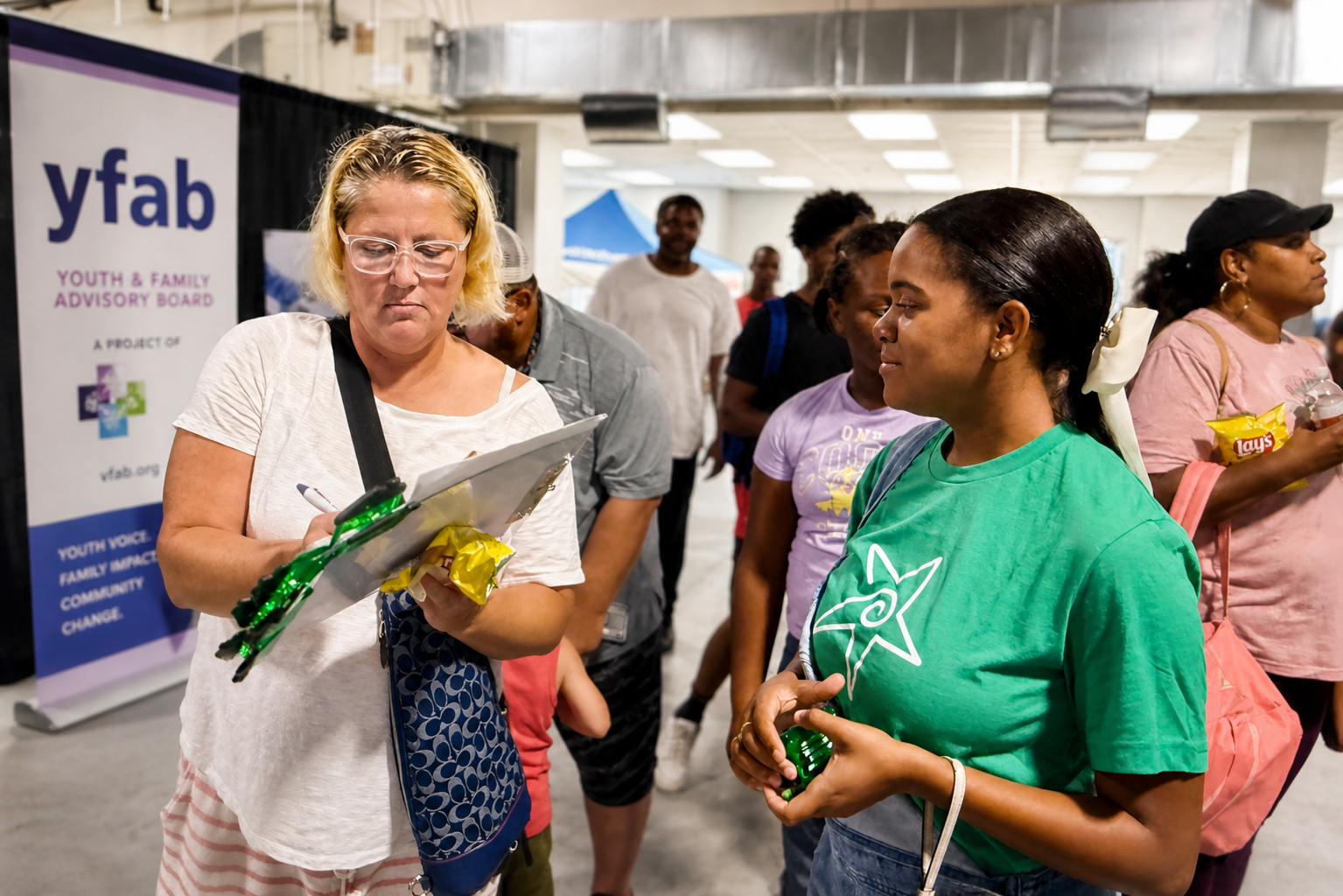 A woman with blonde hair, glasses, and a white shirt signs a clipboard for a woman in a green T-shirt at a community event, with people in line behind them.