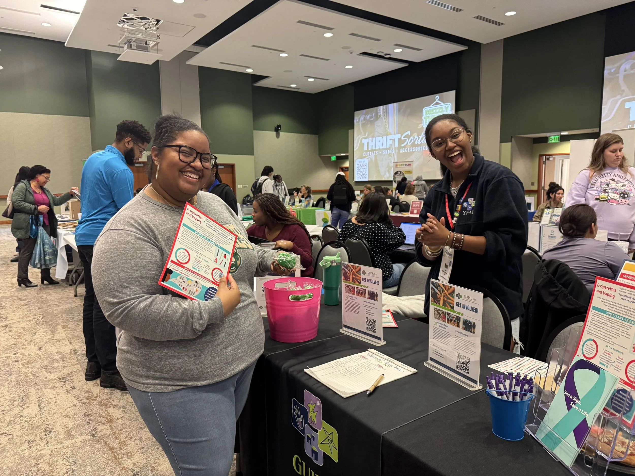 Two women standing at a table in a conference or event hall, smiling at the camera. The table displays informational pamphlets and promotional materials about vaping and e-cigarettes. Other attendees are visible in the background.