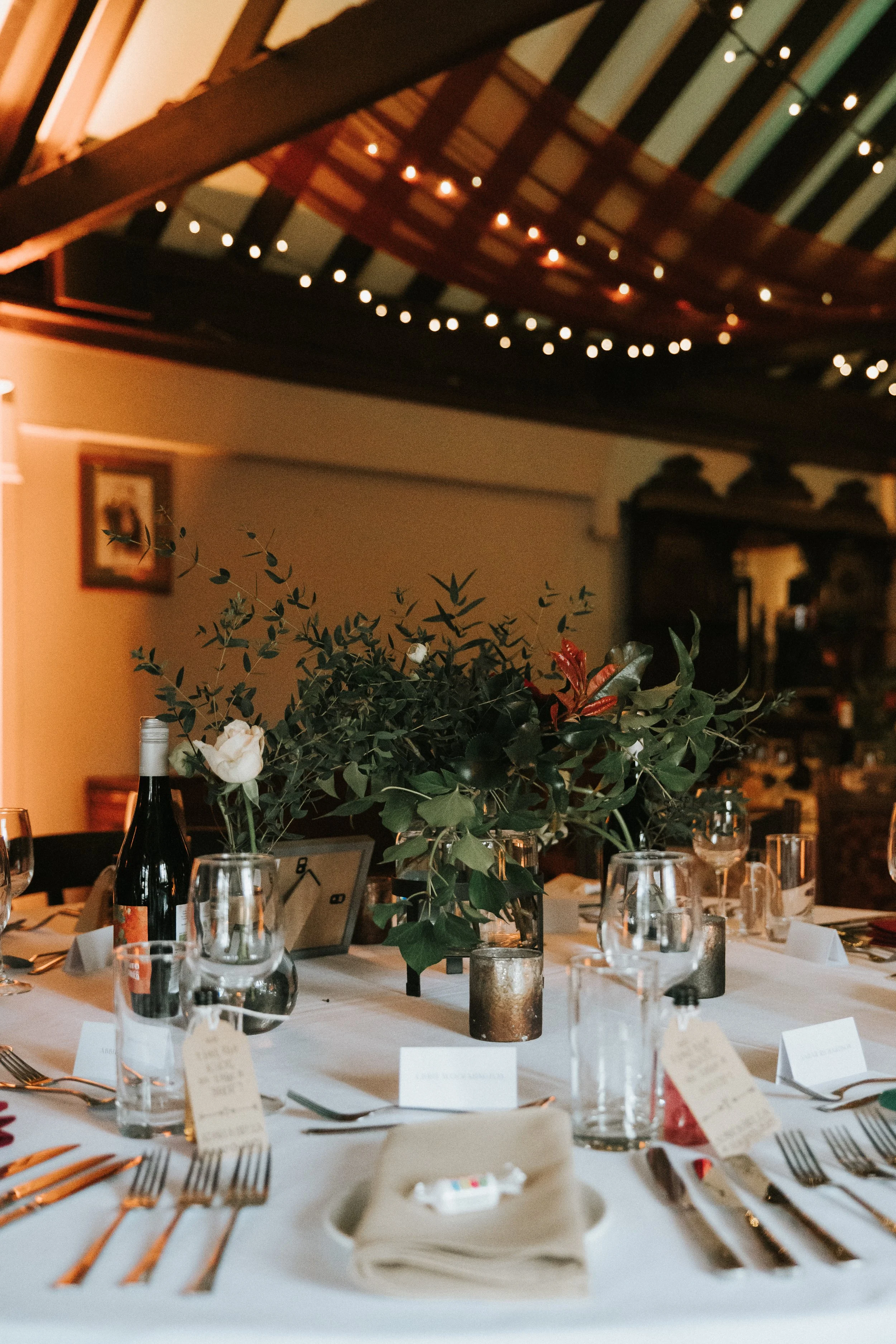 A decorated dining table with a floral centerpiece, wine bottles, glasses, and place cards, set in a warmly lit room with string lights and wooden beams overhead.