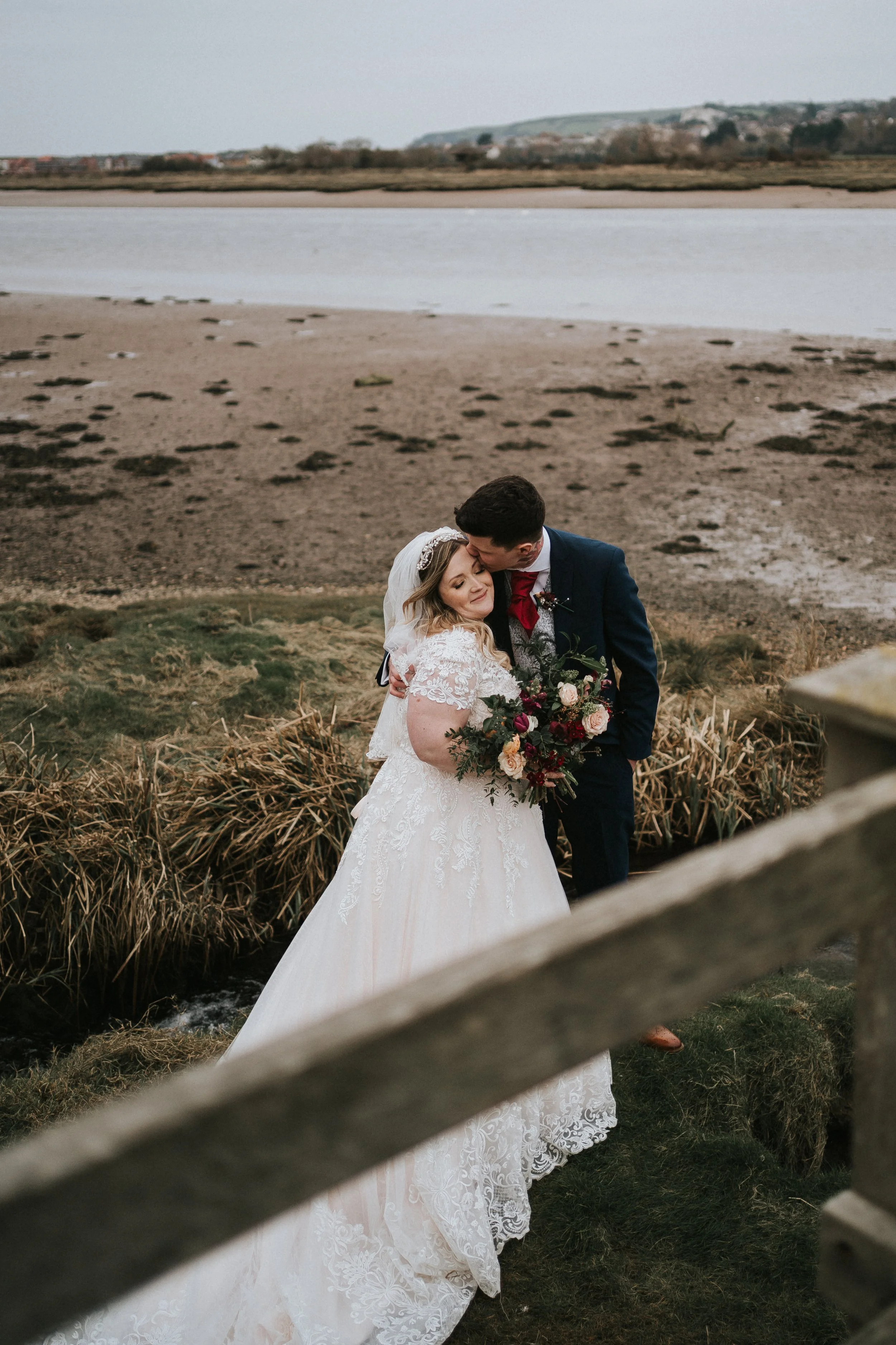 A bride and groom embrace outdoors near a river, with landscape and wetlands in the background. The bride wears a lace wedding dress and veil, holding a bouquet of flowers. The groom is in a dark suit with a red bow tie.