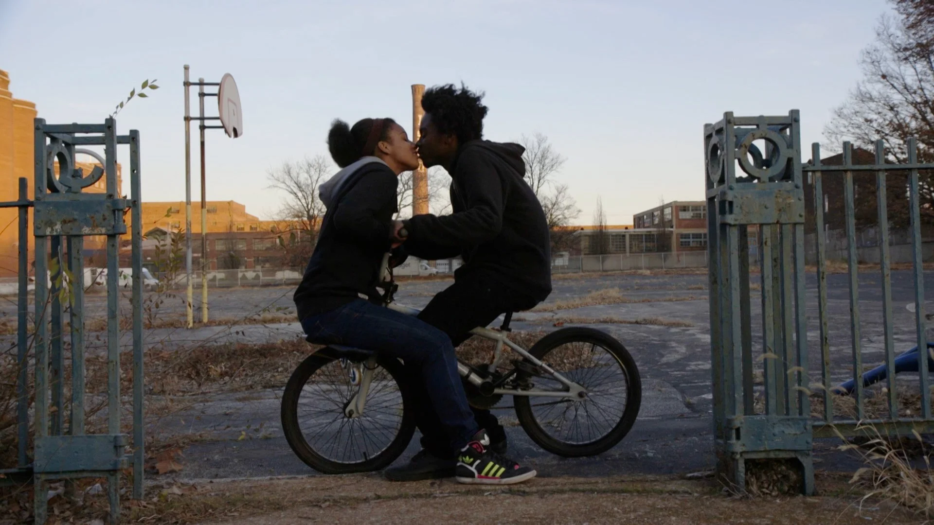 A couple kissing while sitting on a bicycle in an empty playground with a basketball hoop in the background.