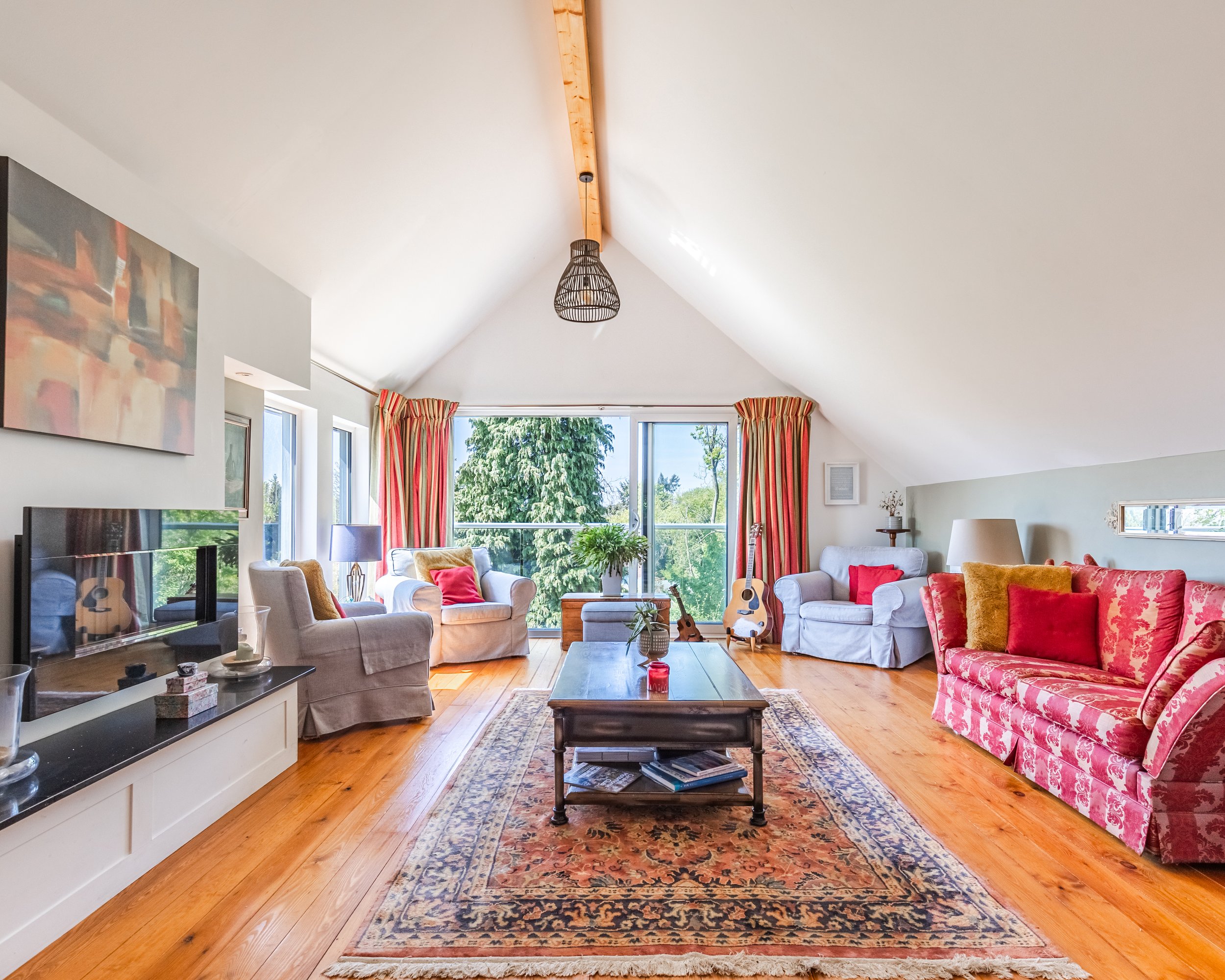 Living room with white ceiling, large windows, red and beige curtains, beige and floral sofas, a wooden coffee table, a patterned rug, a guitar, and Art on the wall.