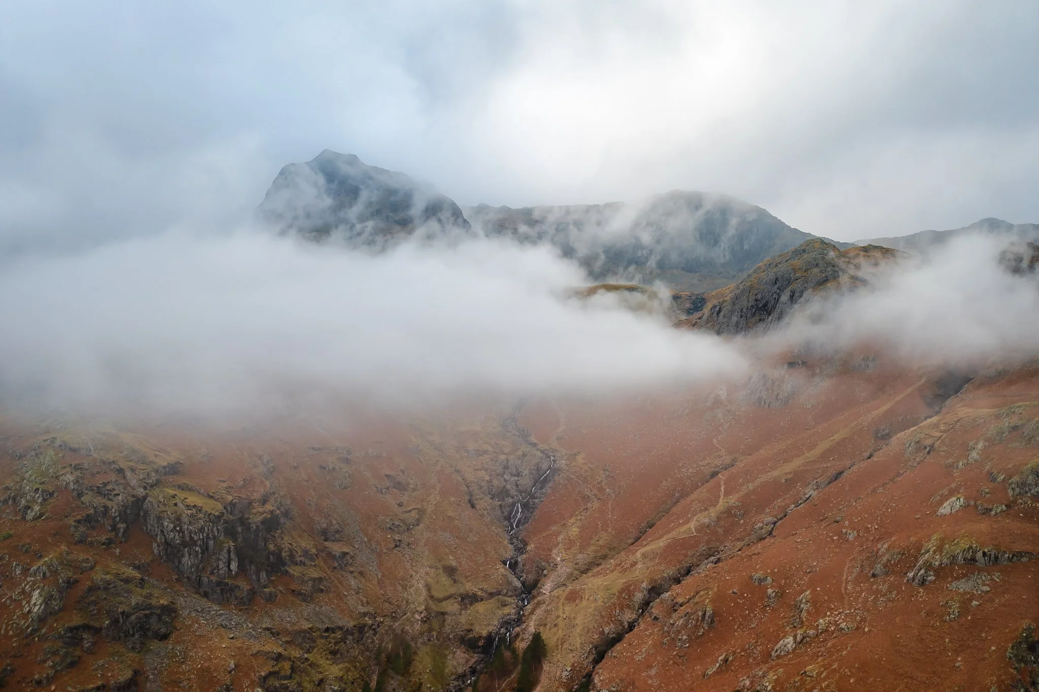 Mountain landscape with clouds and fog, rocky hills, and a small stream in the valley.