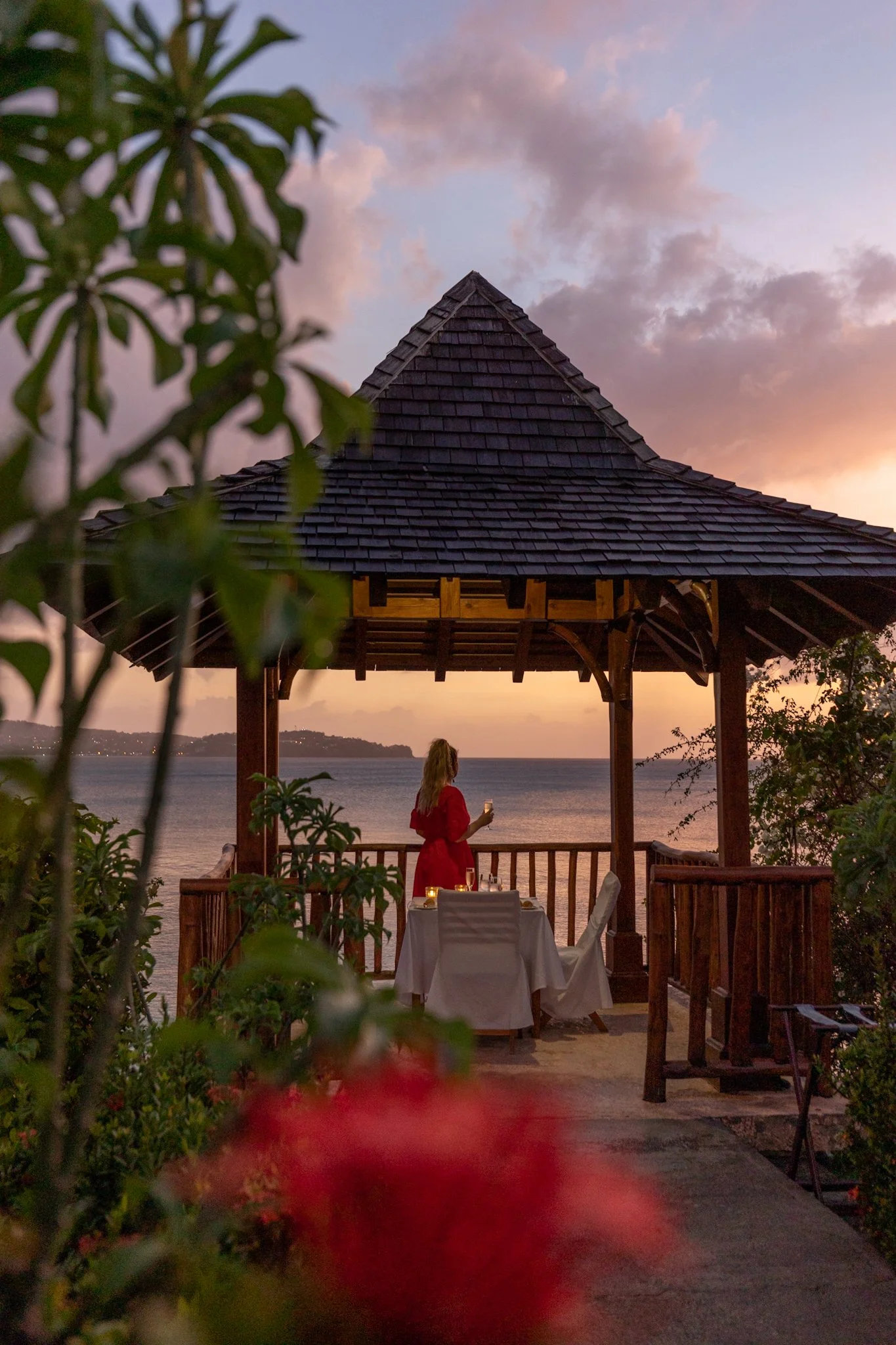 A woman in a red dress stands on a wooden balcony overlooking a body of water during sunset. The sky is partly cloudy with pink and purple hues. There is a table set for dining with a white tablecloth and a candle on it. The area is partially obscure