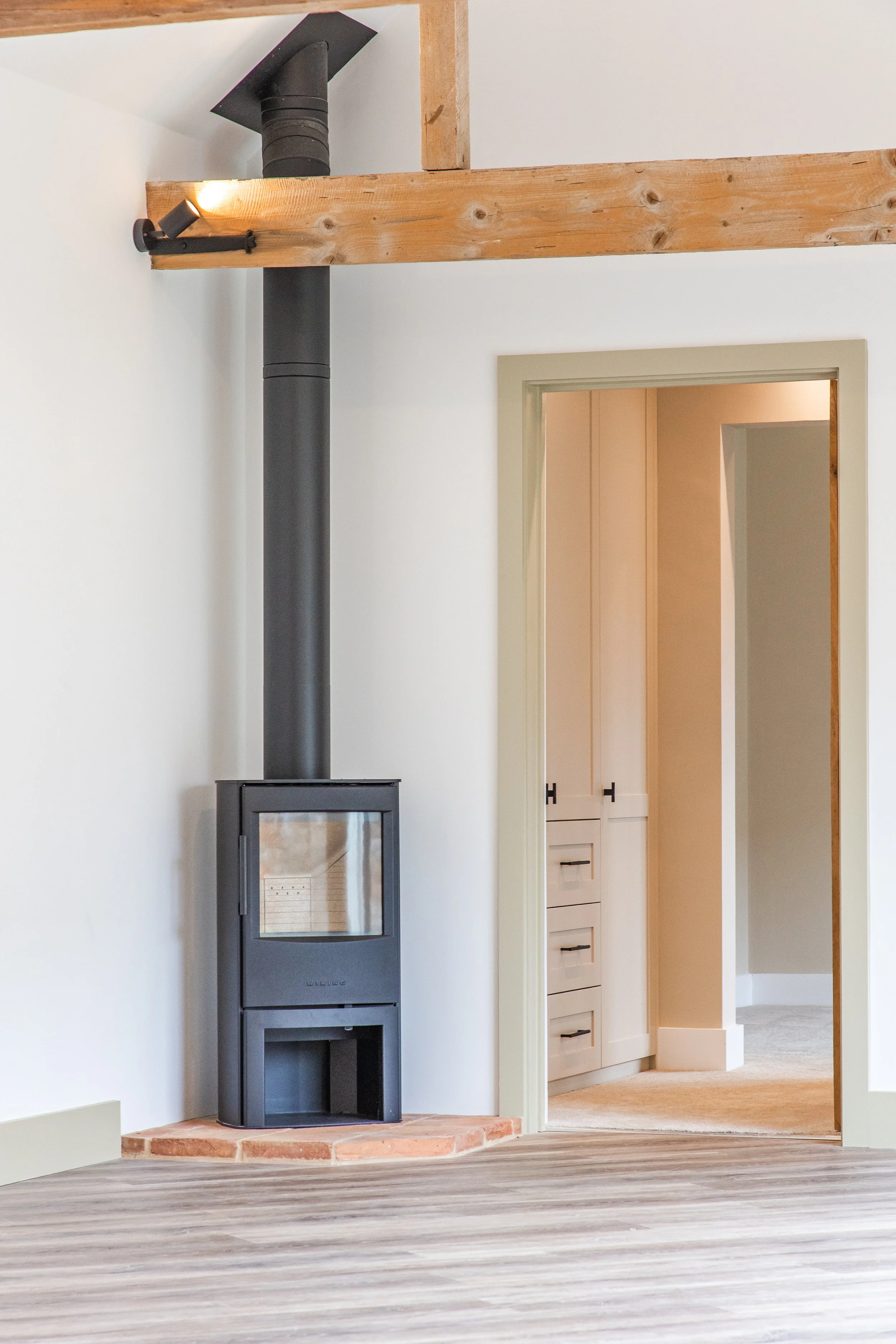 Interior of a house with a modern, black wood stove on a brick platform, wooden beams, and a doorway leading to a room with white cabinets and beige walls.