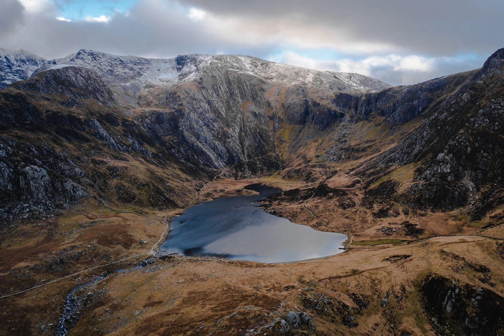 Scenic mountain landscape with snow-capped peaks, rugged cliffs, a lake in the foreground, and cloudy sky.