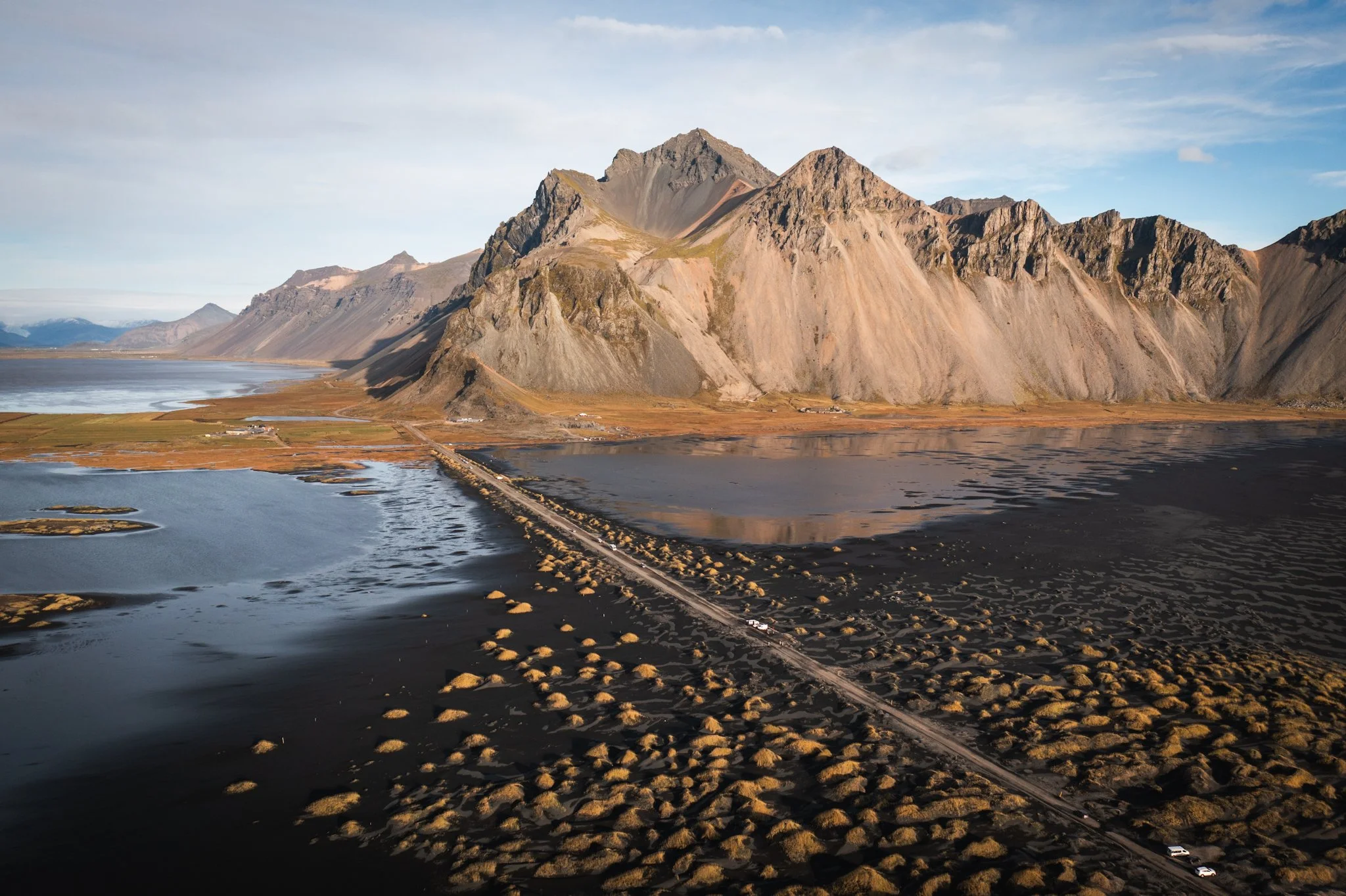 A mountain range with steep steep slopes rising from a flat plain. There is a body of water in the foreground with a road running along its edge, and several small structures are visible near the water.