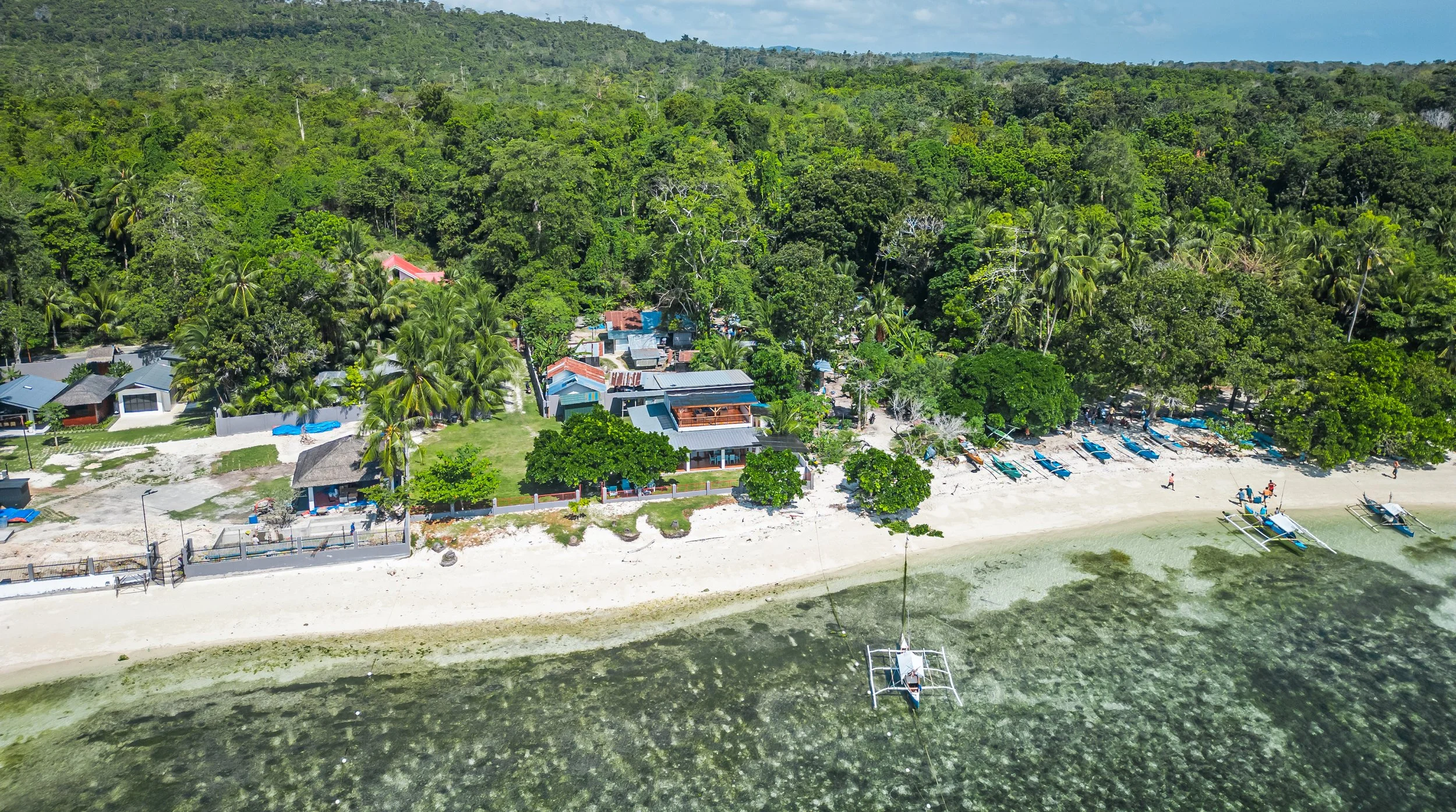 Aerial view of a tropical beach with white sand, boats docked on the shore, dense green forest in the background, and small houses near the beach.