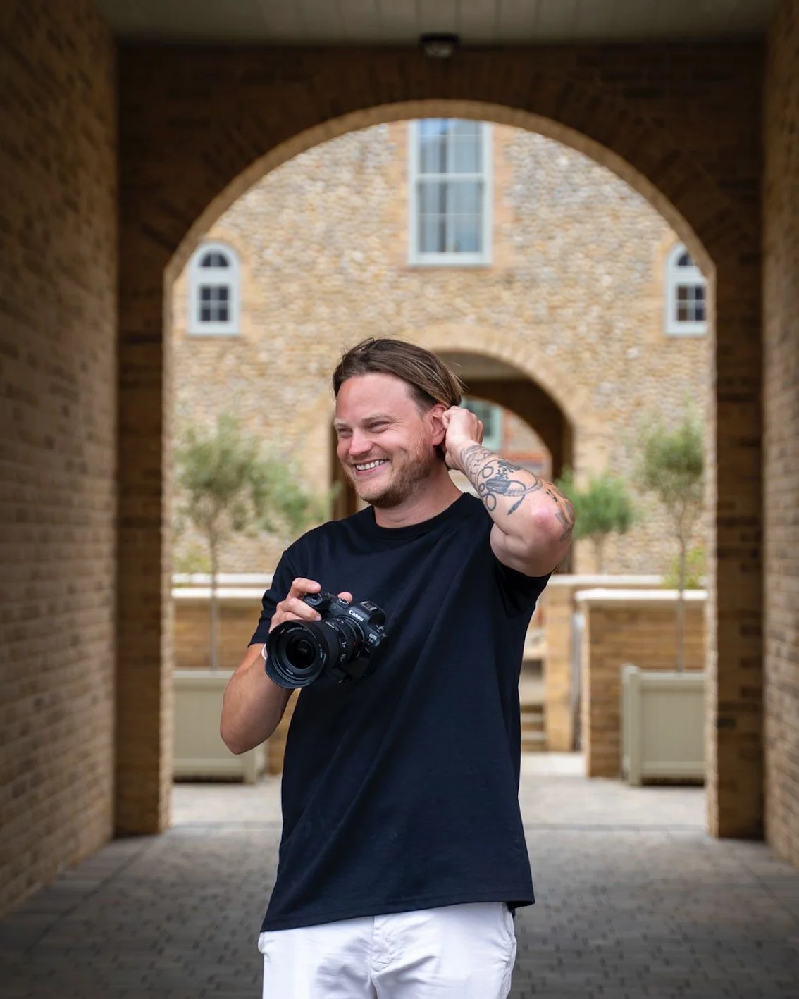 A smiling man with tattoos on his arm, wearing a black t-shirt and white shorts, holding a camera in an urban setting with brick archways and buildings in the background.