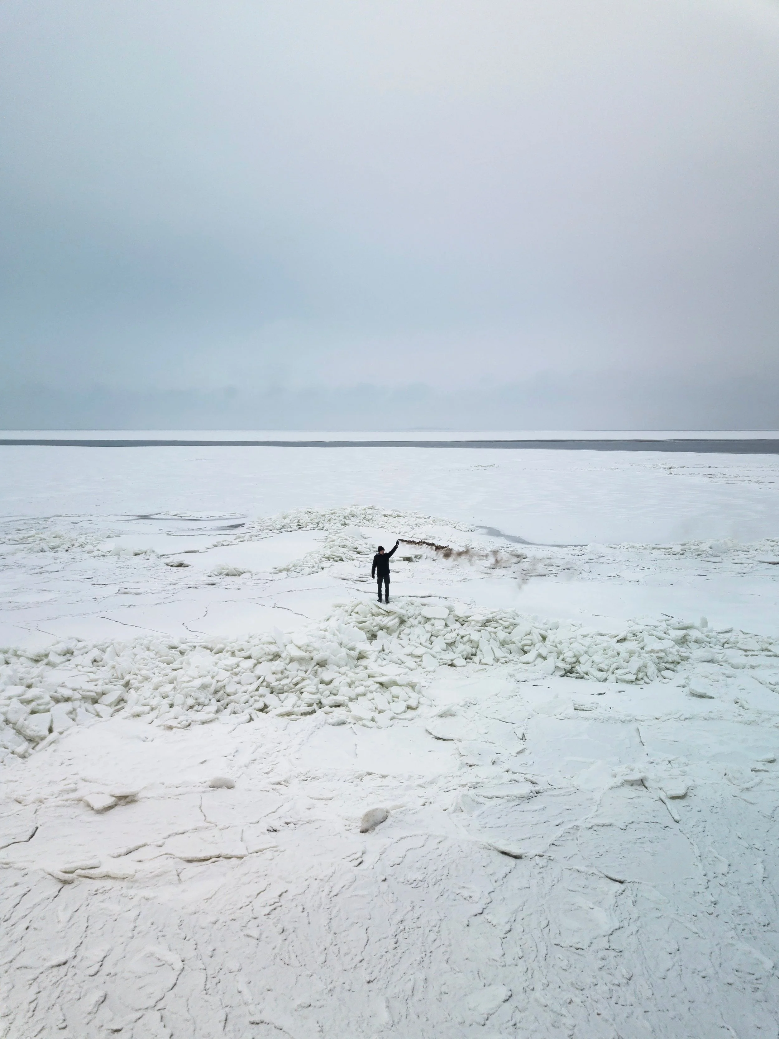 Person standing on icy, snow-covered terrain, with a large body of water or sea in the background and an overcast sky.