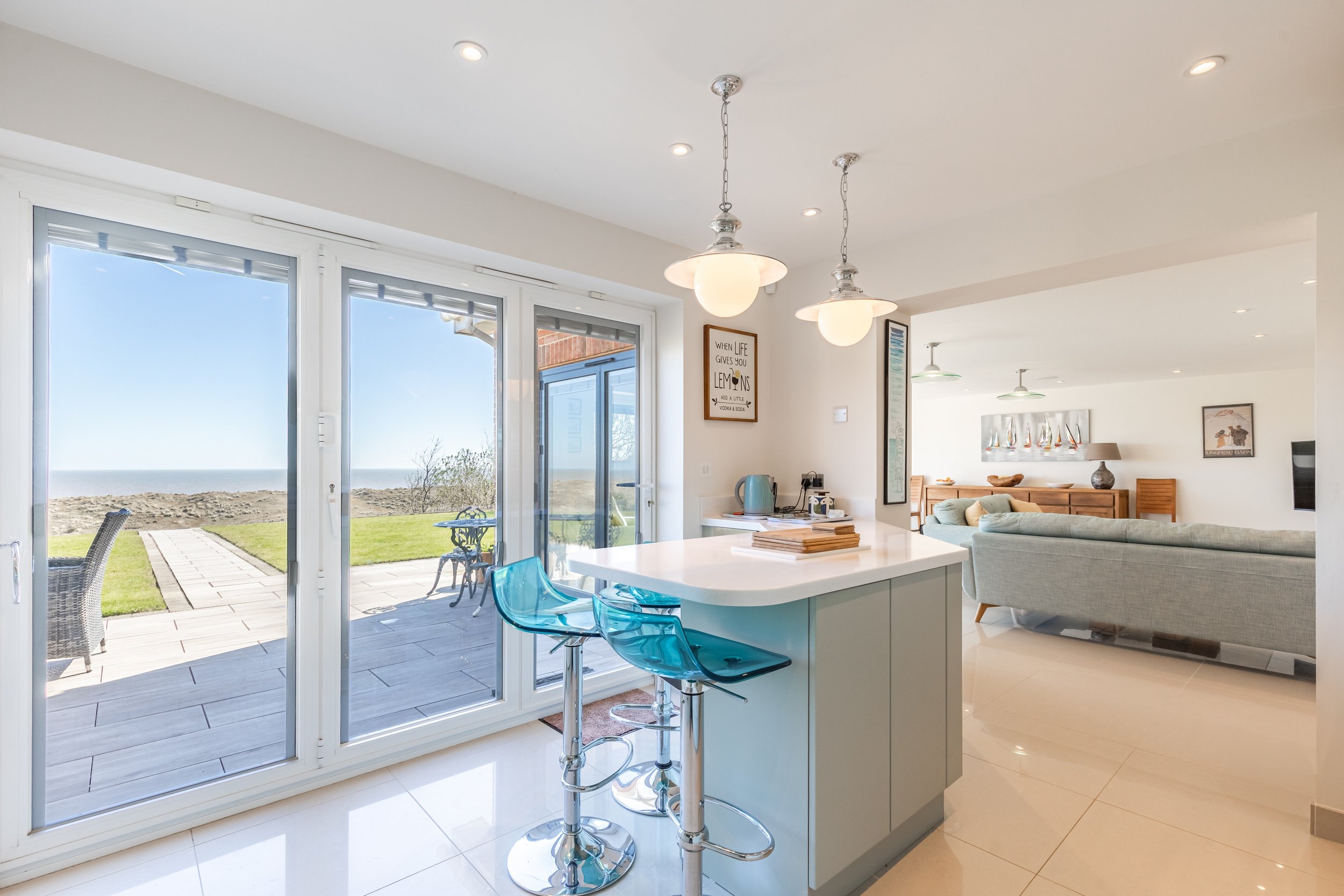 Open concept kitchen and living area with large sliding glass doors leading to an outdoor patio and view of the beach and ocean beyond, decorated with modern furniture and artwork.
