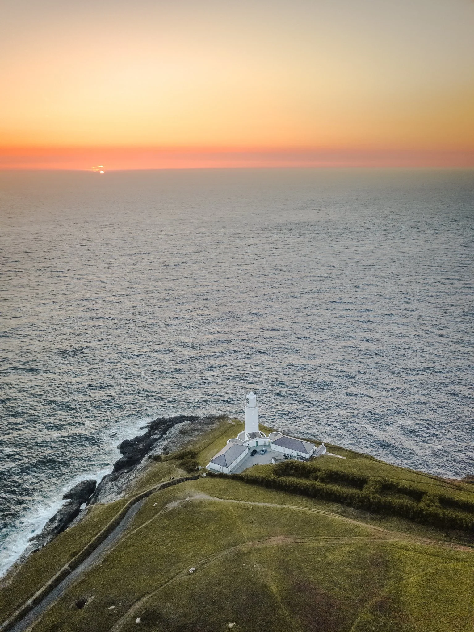 A lighthouse on a rocky coastline with a white building, overlooking the ocean at sunset, with a colorful sky and distant horizon.