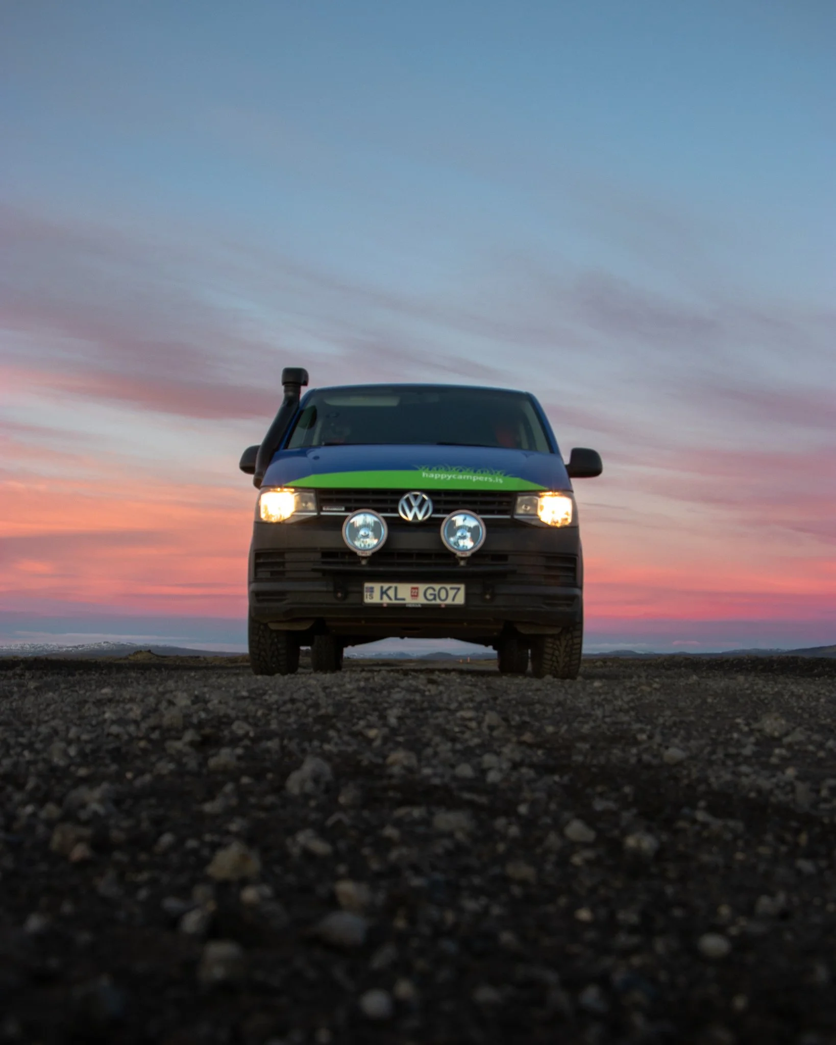 Front view of a Volkswagen van with a green stripe and logo, parked on gravel ground with a sunset sky in the background.