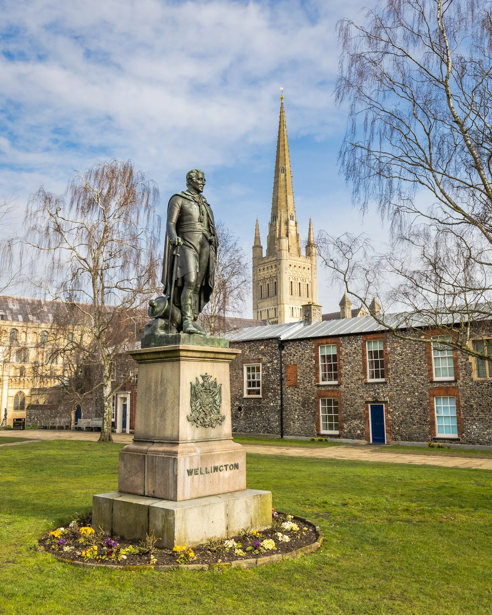 A statue of William Wellington in a park with a flower bed at its base, a tower in the background, and leafless trees around.