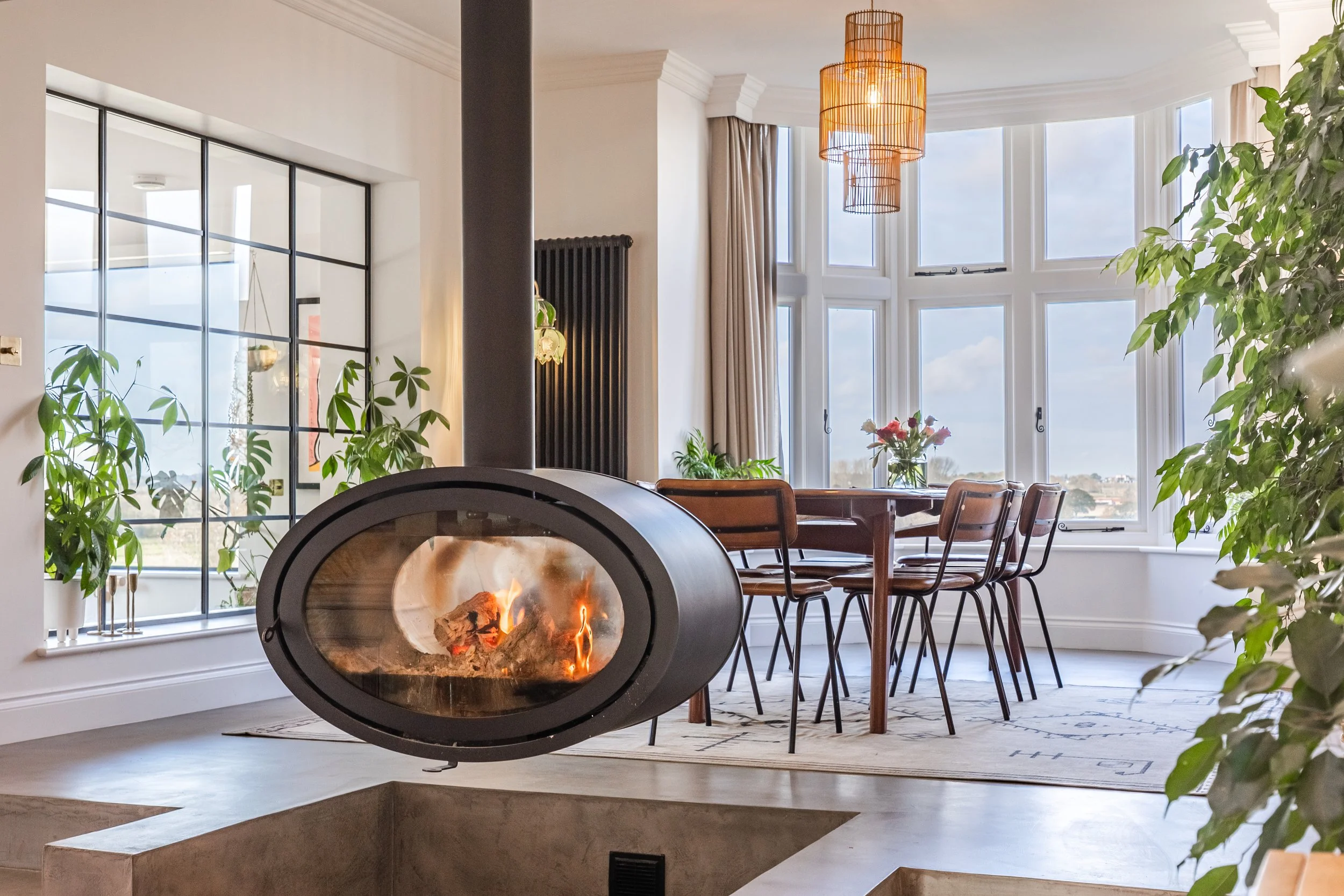A modern living room with a fireplace, a dining table with chairs, large bay windows with curtains, and green houseplants.