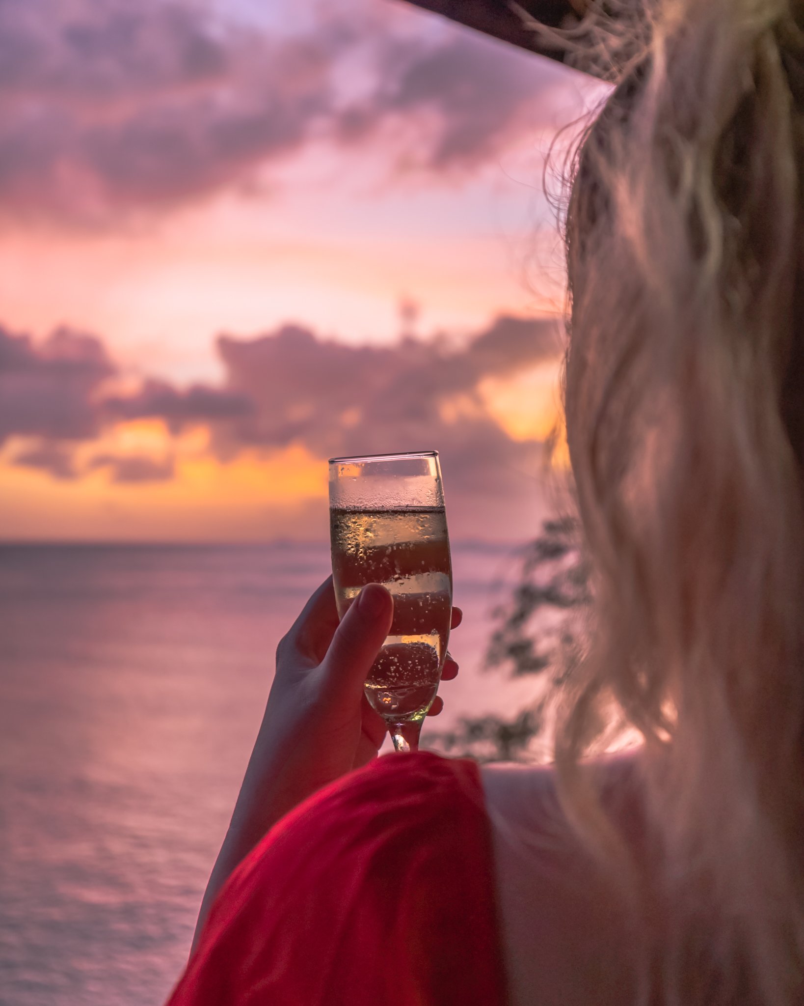 Person holding a glass of champagne or sparkling wine during sunset by the water with clouds and sky in the background.