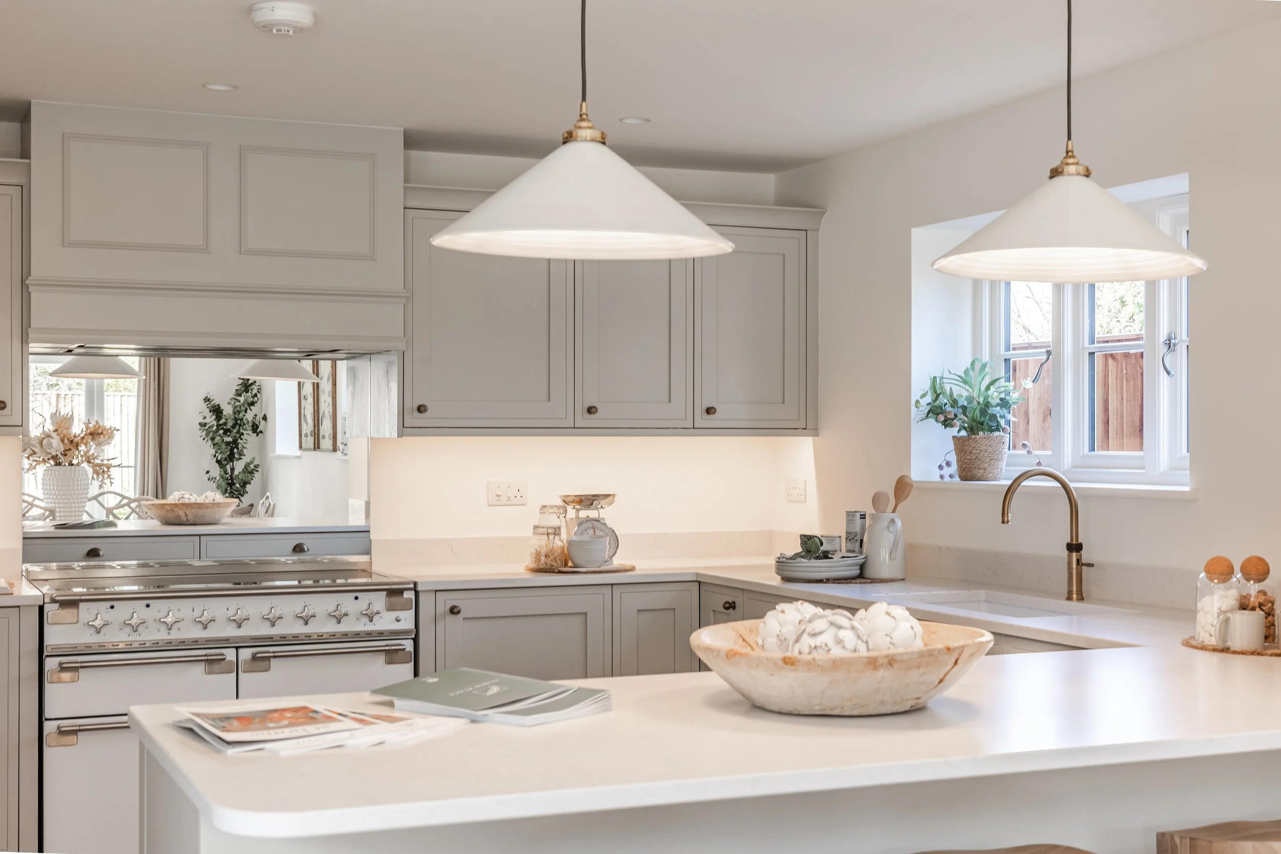 Bright, modern kitchen with white countertops, light gray cabinetry, and large windows letting in natural light. Decor includes plants, a bowl of cotton, and kitchen utensils.