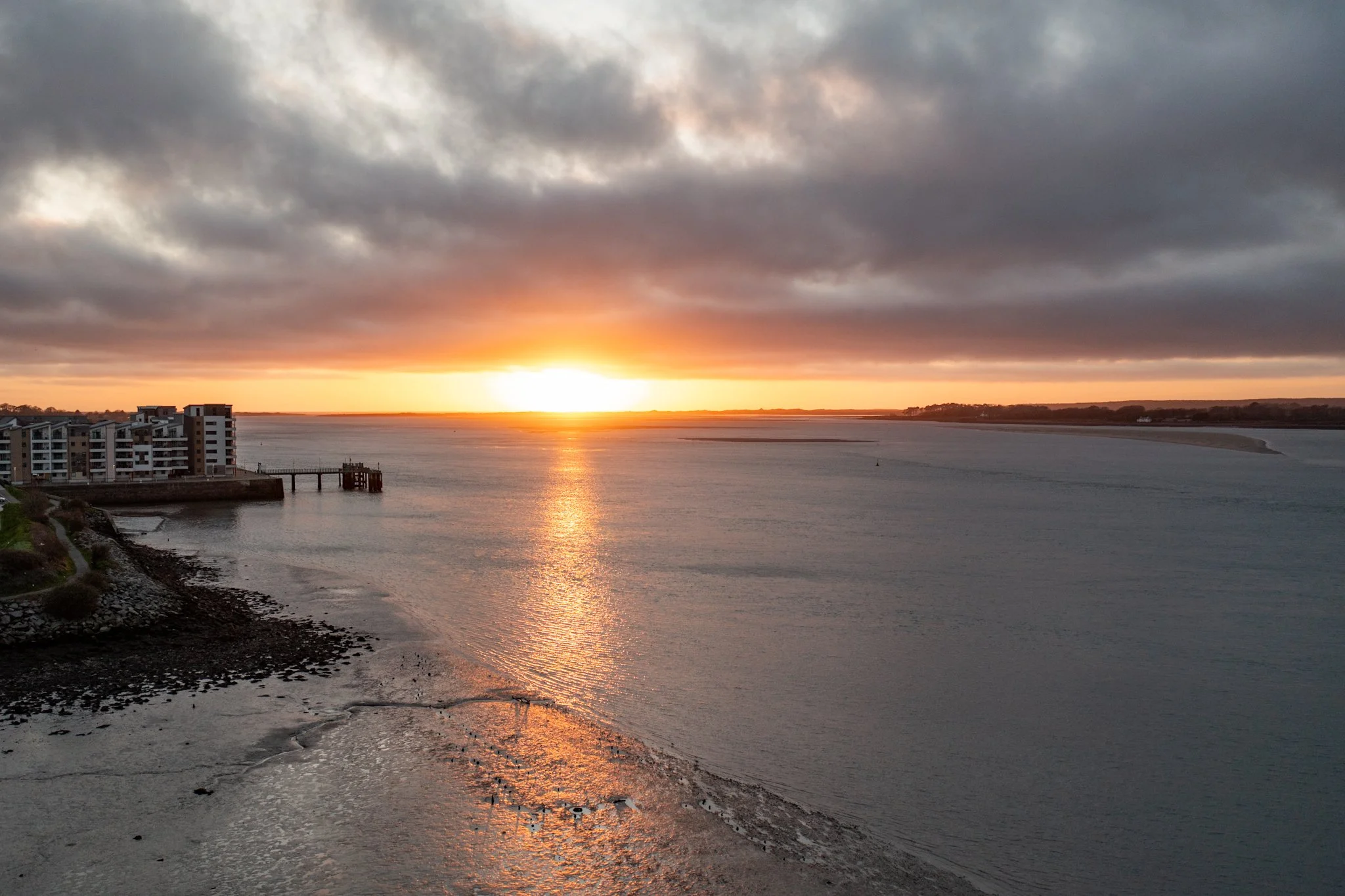 Sunset over a body of water with clouds covered sky. Residential buildings are on the left, and a small pier extends into the water. The shoreline is rocky and muddy in the foreground.
