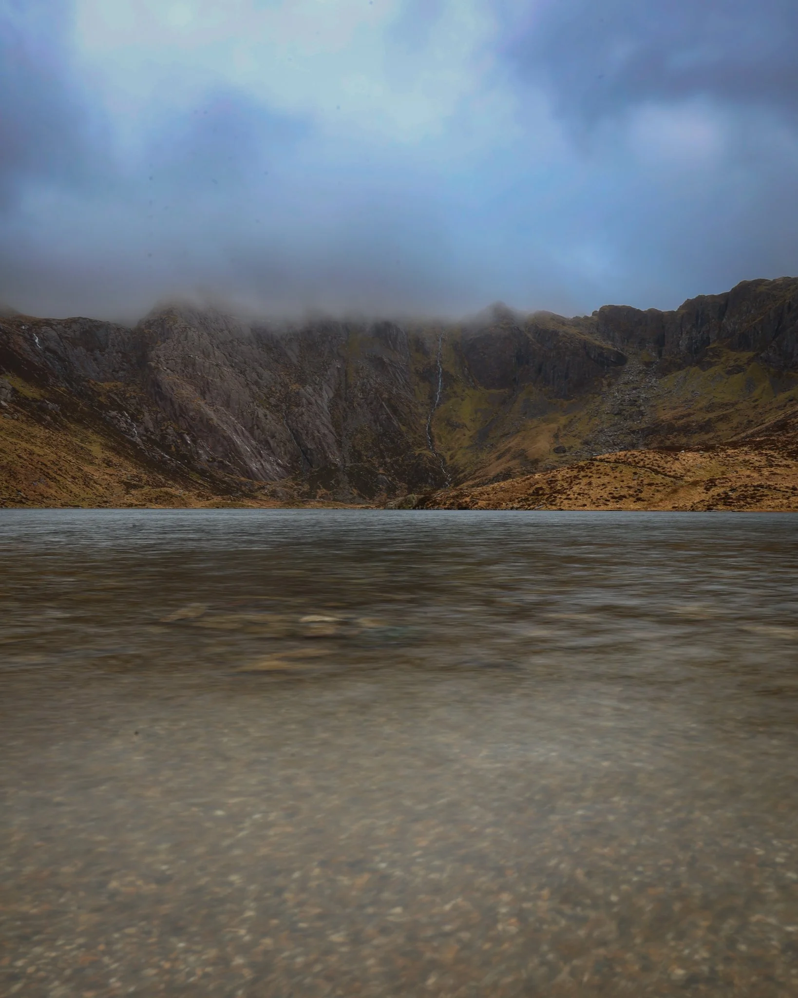 A landscape with a body of water in the foreground, steep rocky mountains with patches of moss and small waterfalls in the middle ground, and a cloudy sky with fog covering the mountain tops.