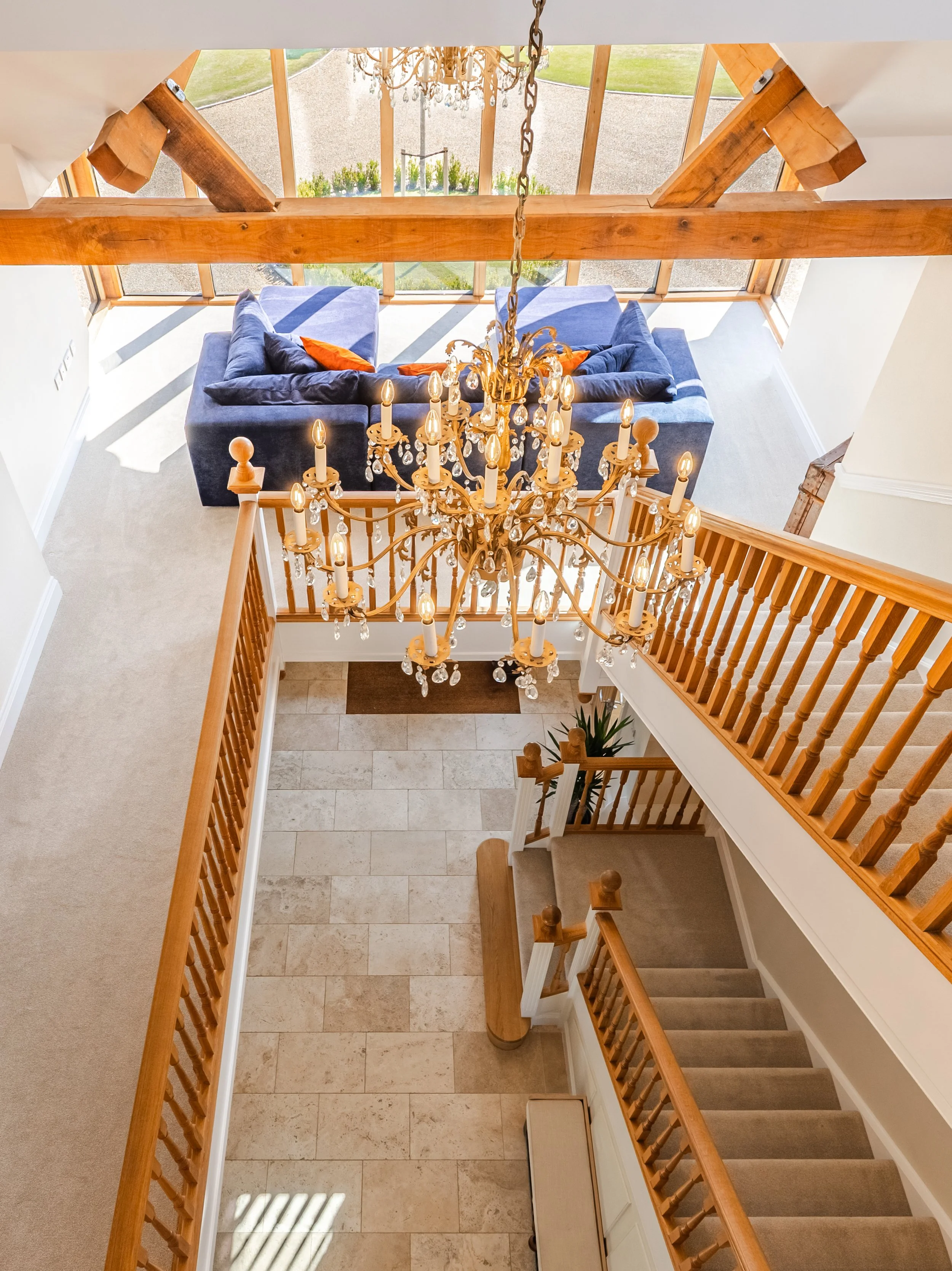 View from the second floor of a house, looking down at the staircase and chandelier, with a sitting area and sliding glass doors leading outside to a garden with grass and gravel.