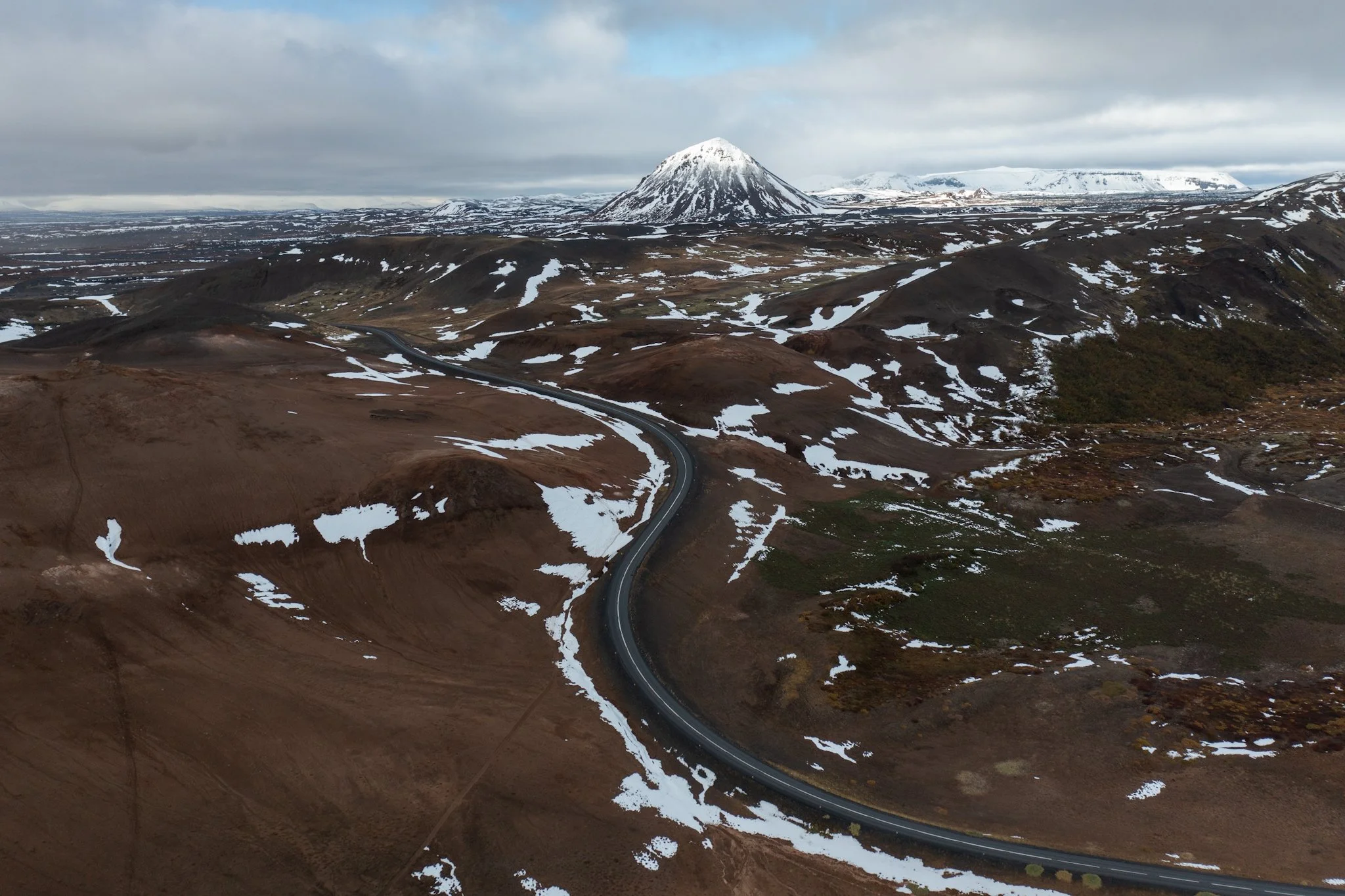 A mountainous landscape with a prominent snow-capped cone-shaped volcano in the background, rugged terrain with patches of snow, and a winding road through the brownish ground under a cloudy sky.
