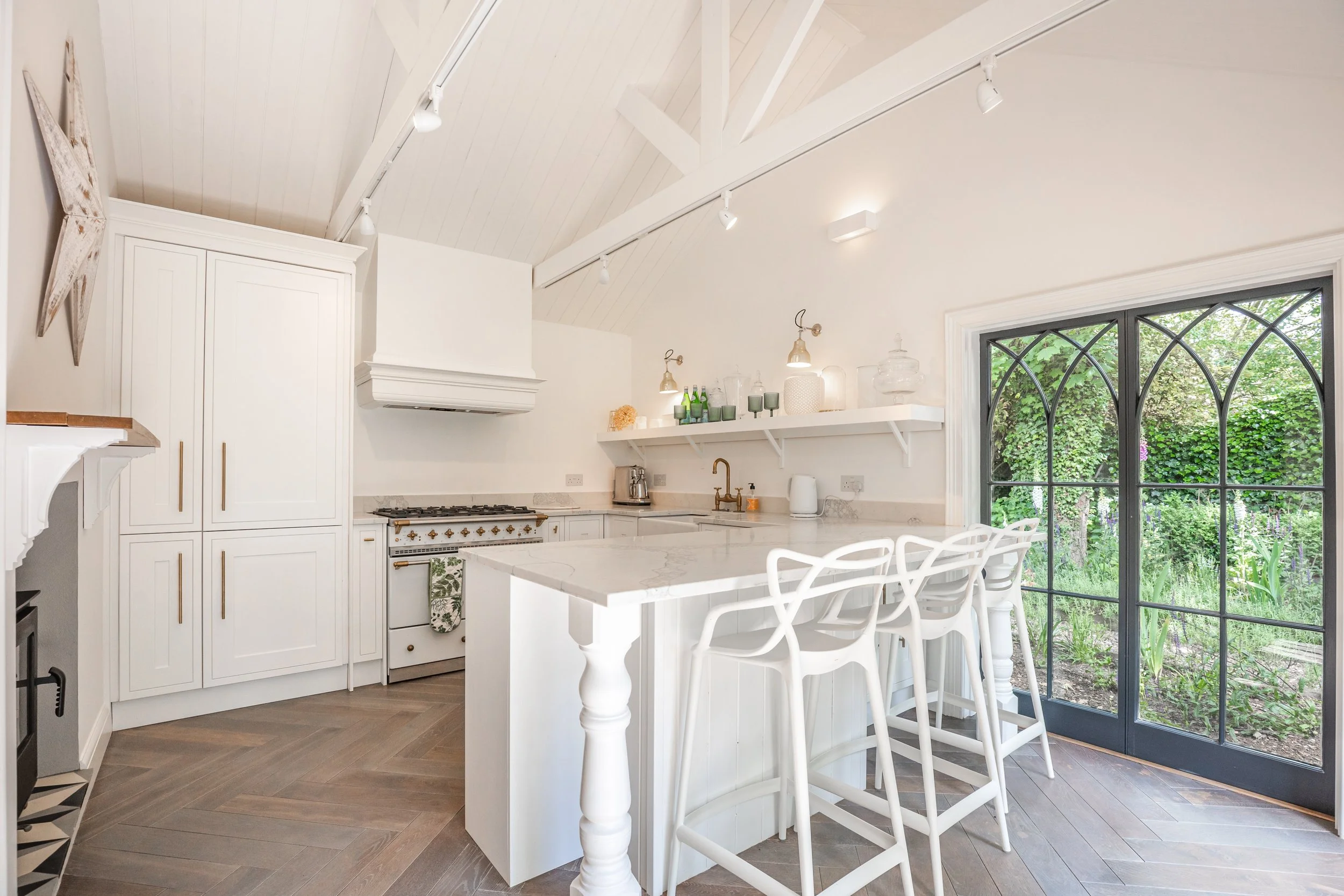 A bright, white kitchen featuring a white island with decorative legs and a marble countertop, white cabinets, and open shelving with decorative glass containers and green bottles. There is a large window with black framing showing green garden outsi