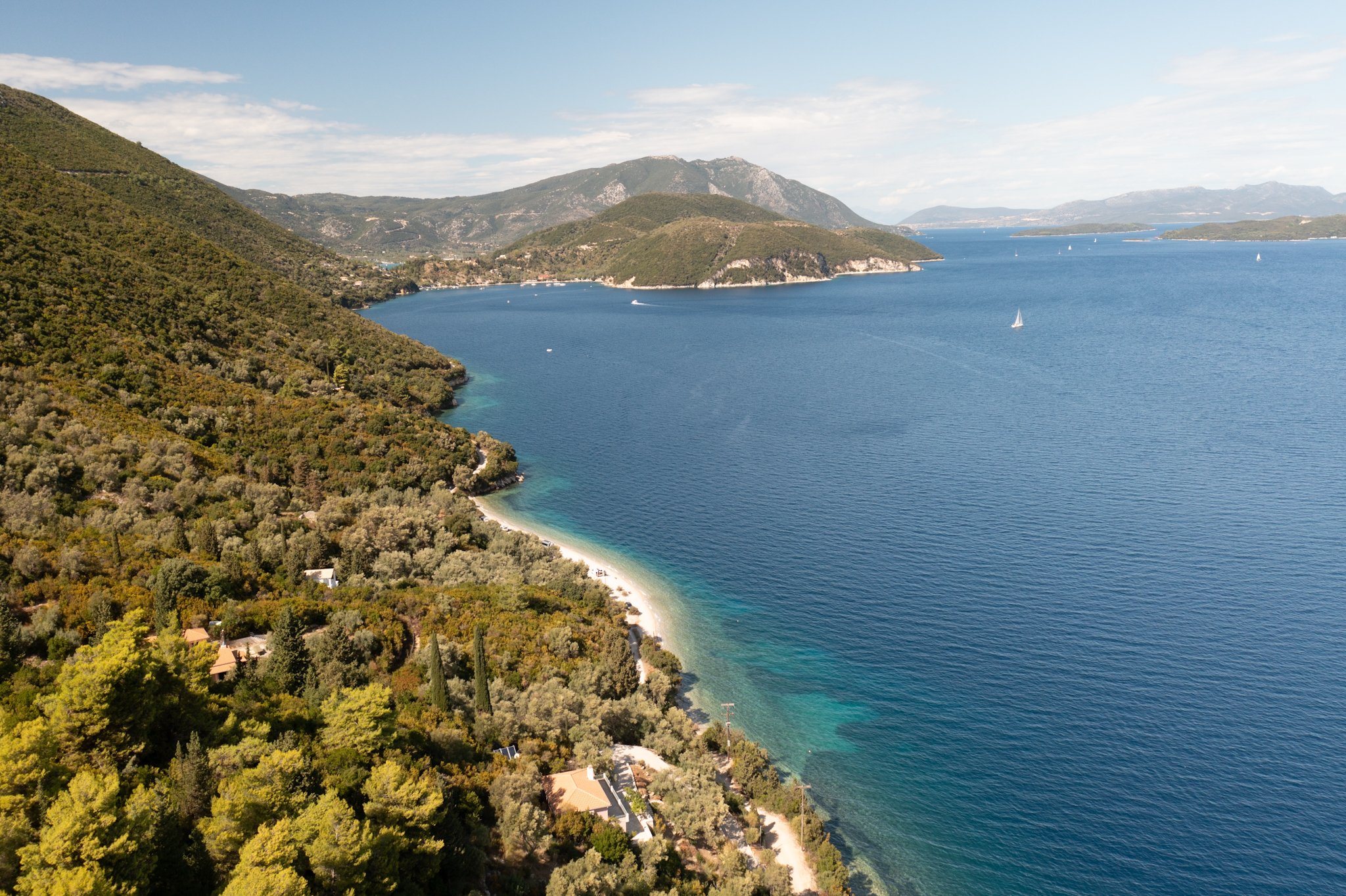 Aerial view of a lake with sailboats, surrounded by lush green hills and mountains under a partly cloudy sky.