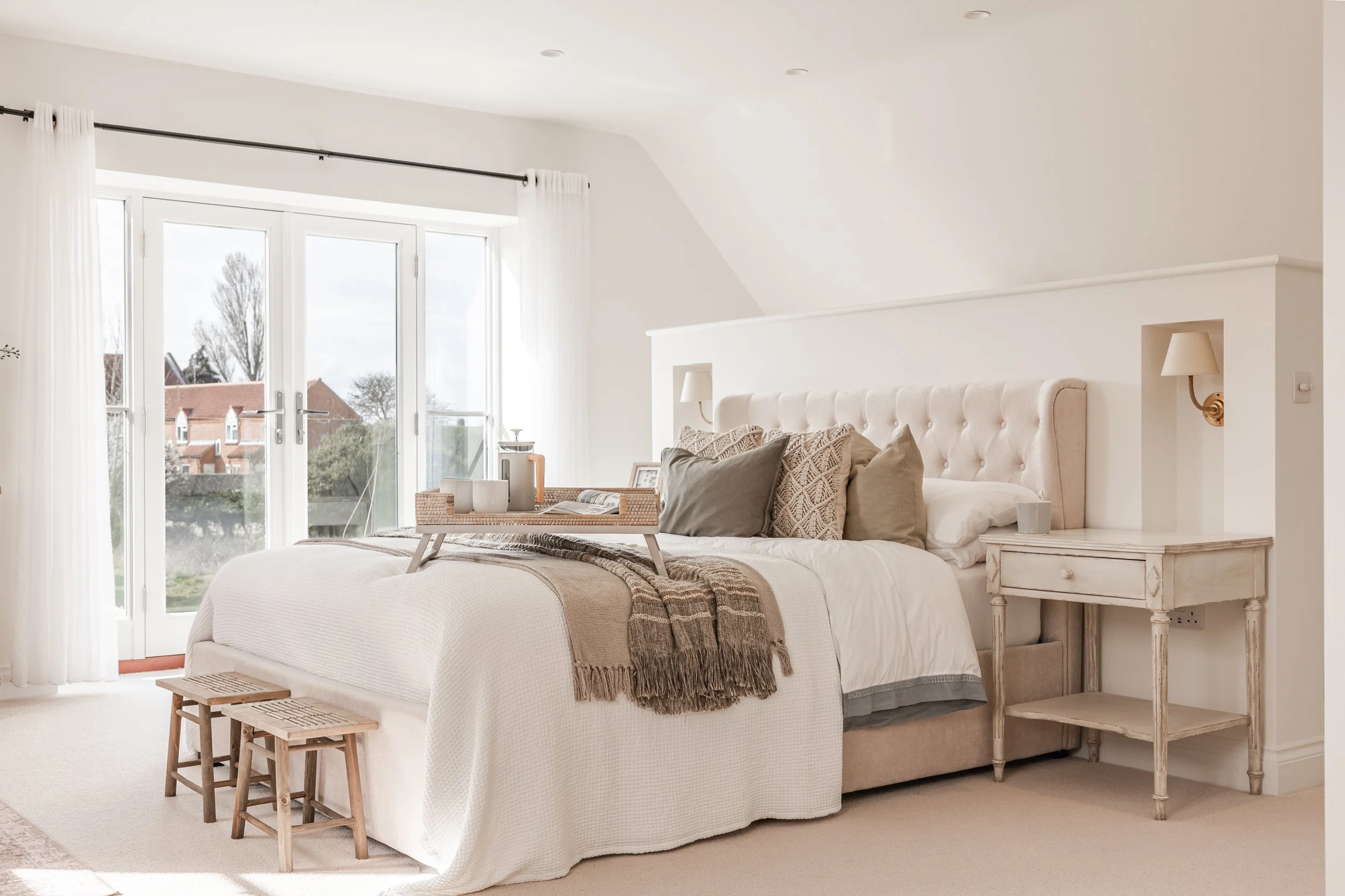 A cozy bedroom with a large bed covered in white bedding and decorative pillows. There is a wooden tray with cups on top of the bed. The room has a white color scheme with a large window/door with white curtains, letting in natural light and showing 