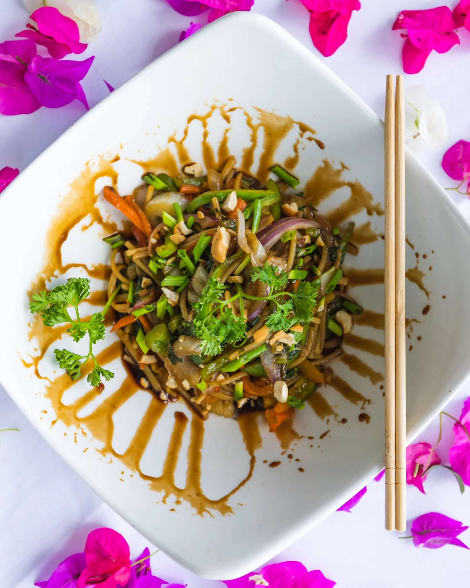 A partially eaten Asian noodle dish with vegetables and sauce on a white square plate, garnished with cilantro and chopsticks on the side, surrounded by pink bougainvillea flowers.