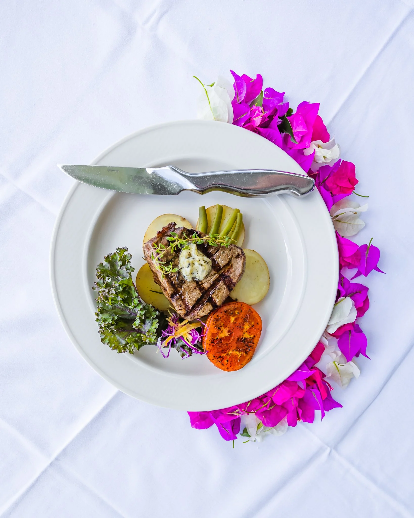 Plate with grilled steak, roasted potato slices, green beans, grilled tomato, and garnishes, surrounded by purple and white flowers on white tablecloth.