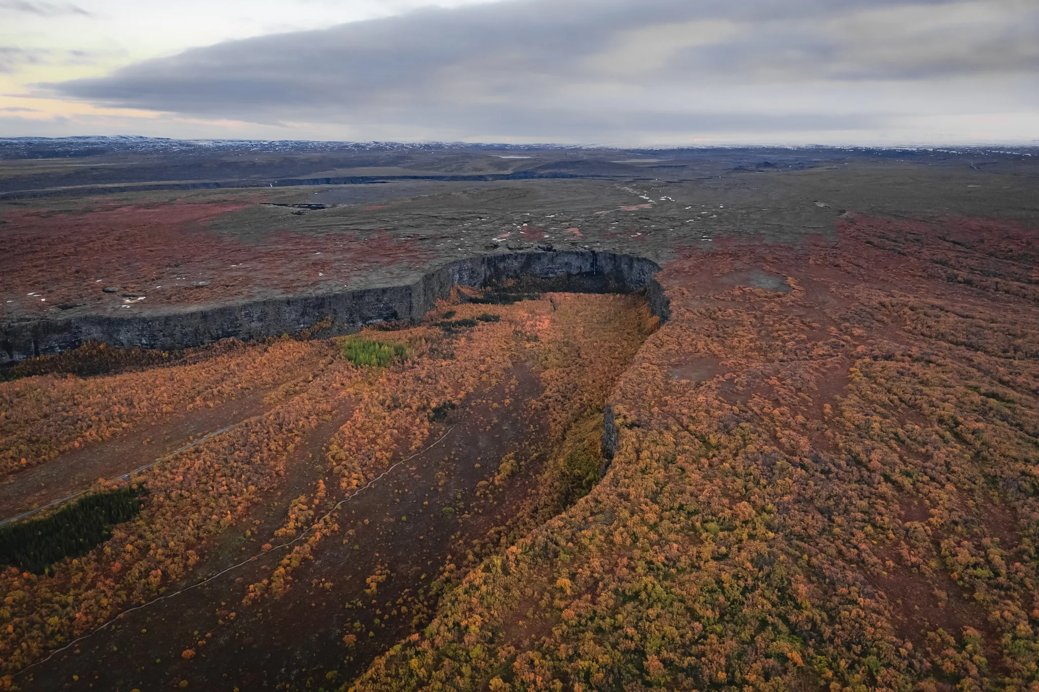 An aerial view of a crater surrounded by colorful autumn trees with sparse patches of snow and a distant cloudy sky.