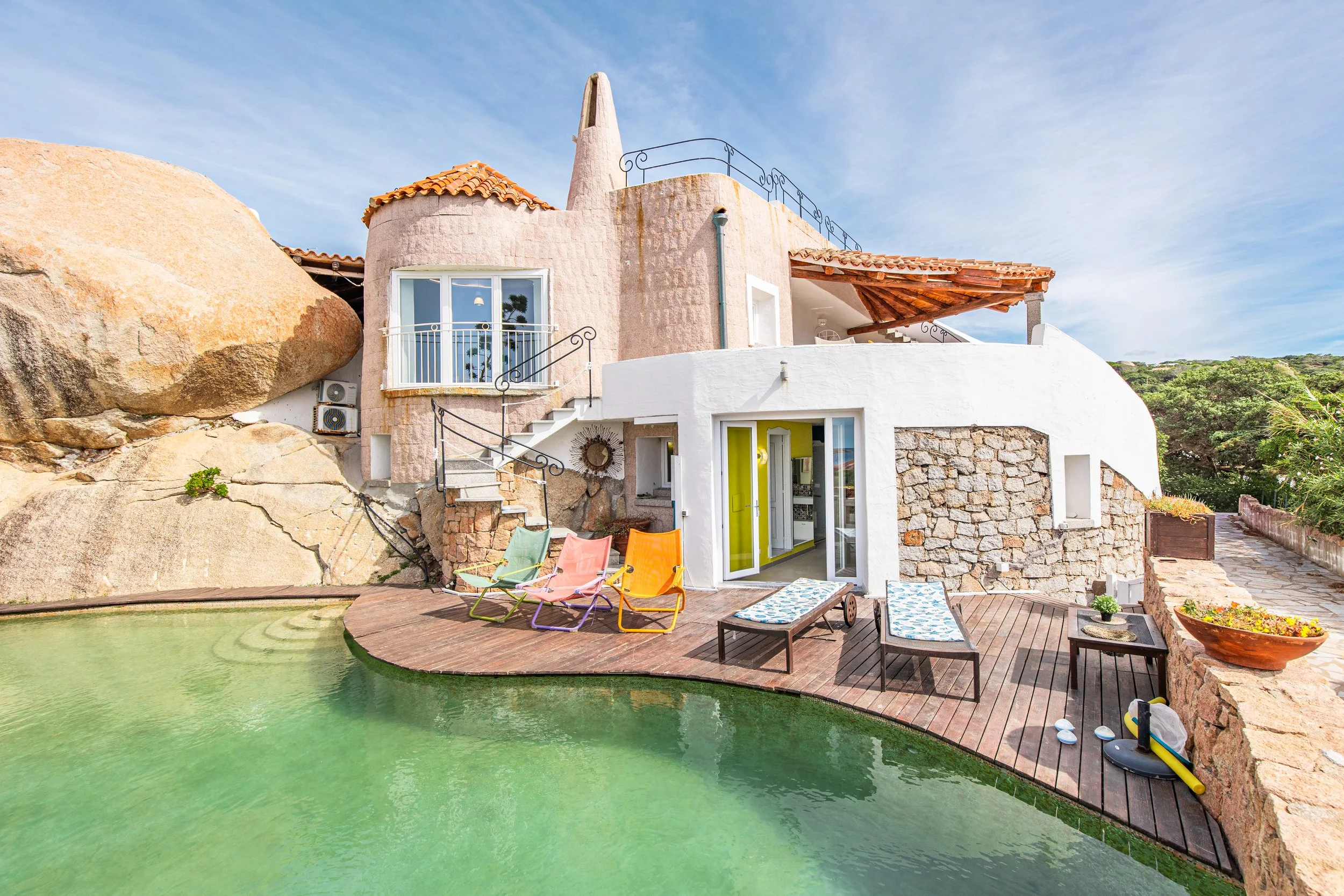 A Sardinian Villa built into large rocks with a swimming pool and colorful lounge chairs outside, under a partly cloudy sky.