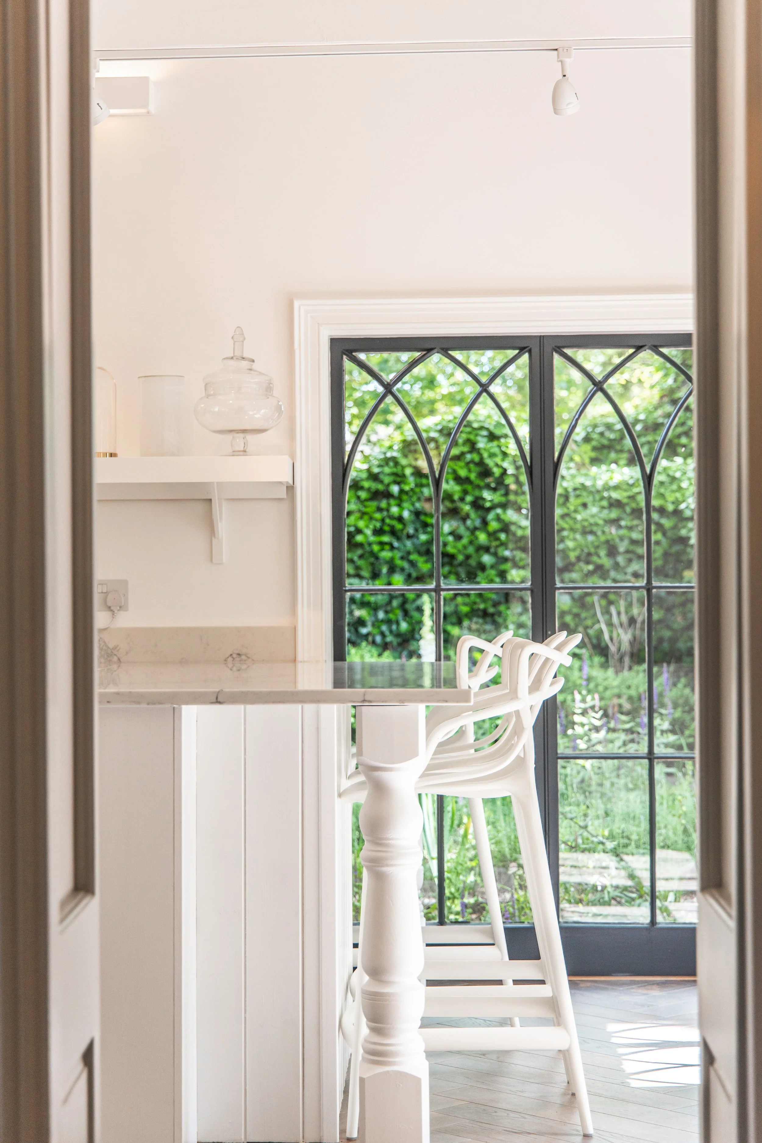View through a doorway into a bright room with a large window showing green bushes outside. Inside, there is a white table with white chairs, a white shelf with decorative glass containers, and a ceiling with white track lighting.
