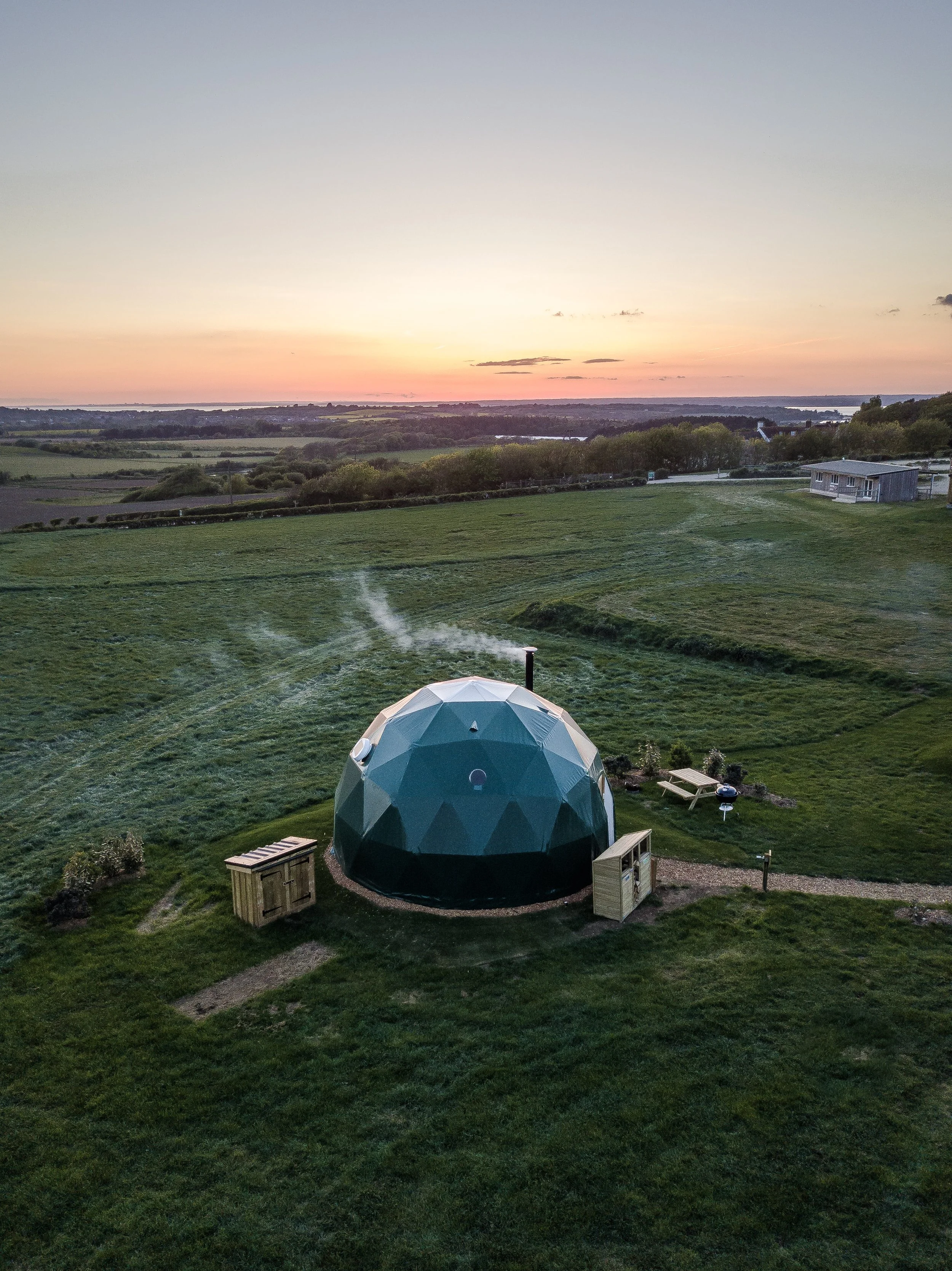 A geodesic dome structure on a grassy landscape during sunset, with a gravel path leading up to it and a small wooden shed nearby.