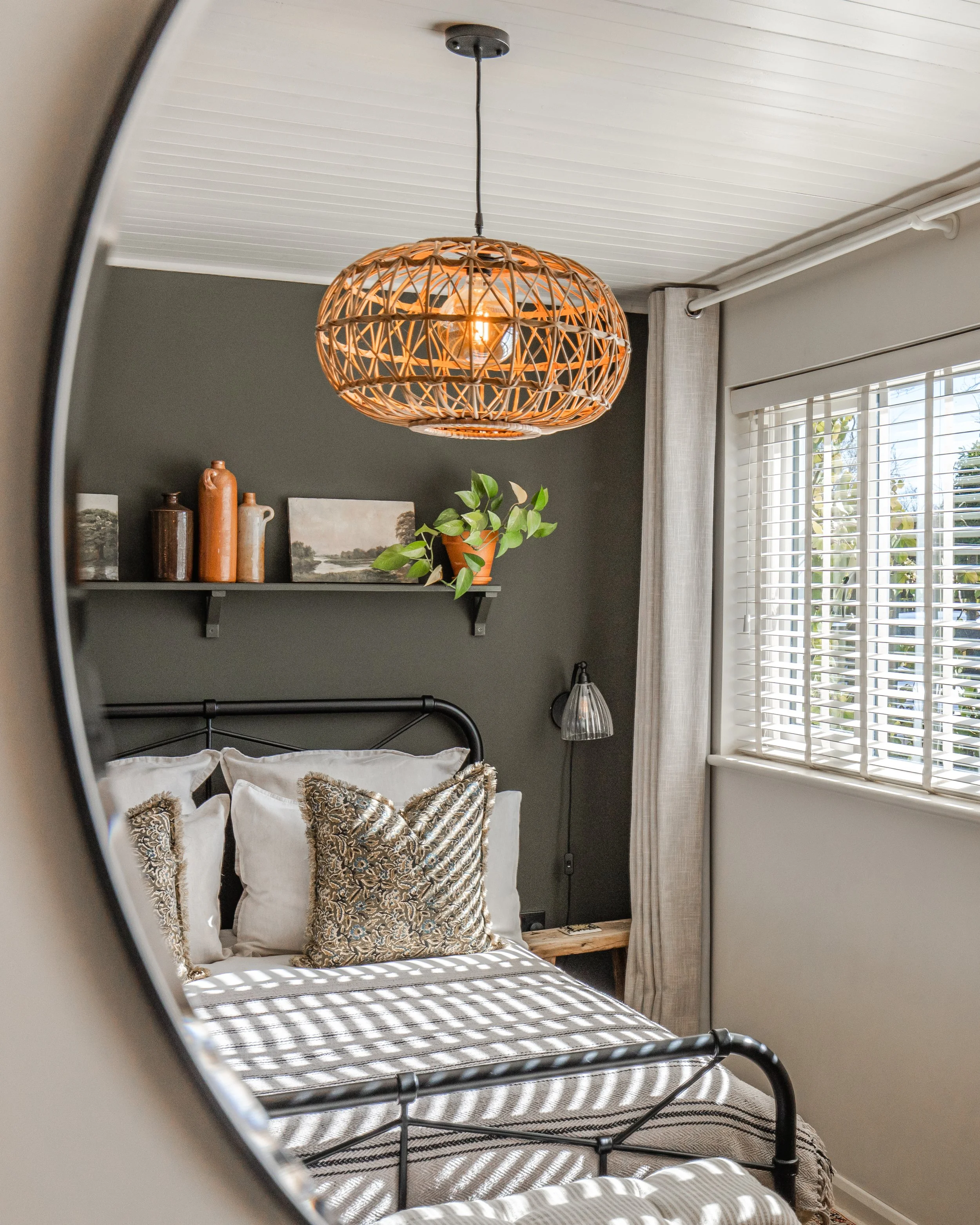 A bedroom featuring a black metal bed frame with a striped bedspread, decorative pillows with animal print, a wooden shelf with patterned vases and a landscape painting, and a window with white blinds and beige curtains. A modern woven pendant light 